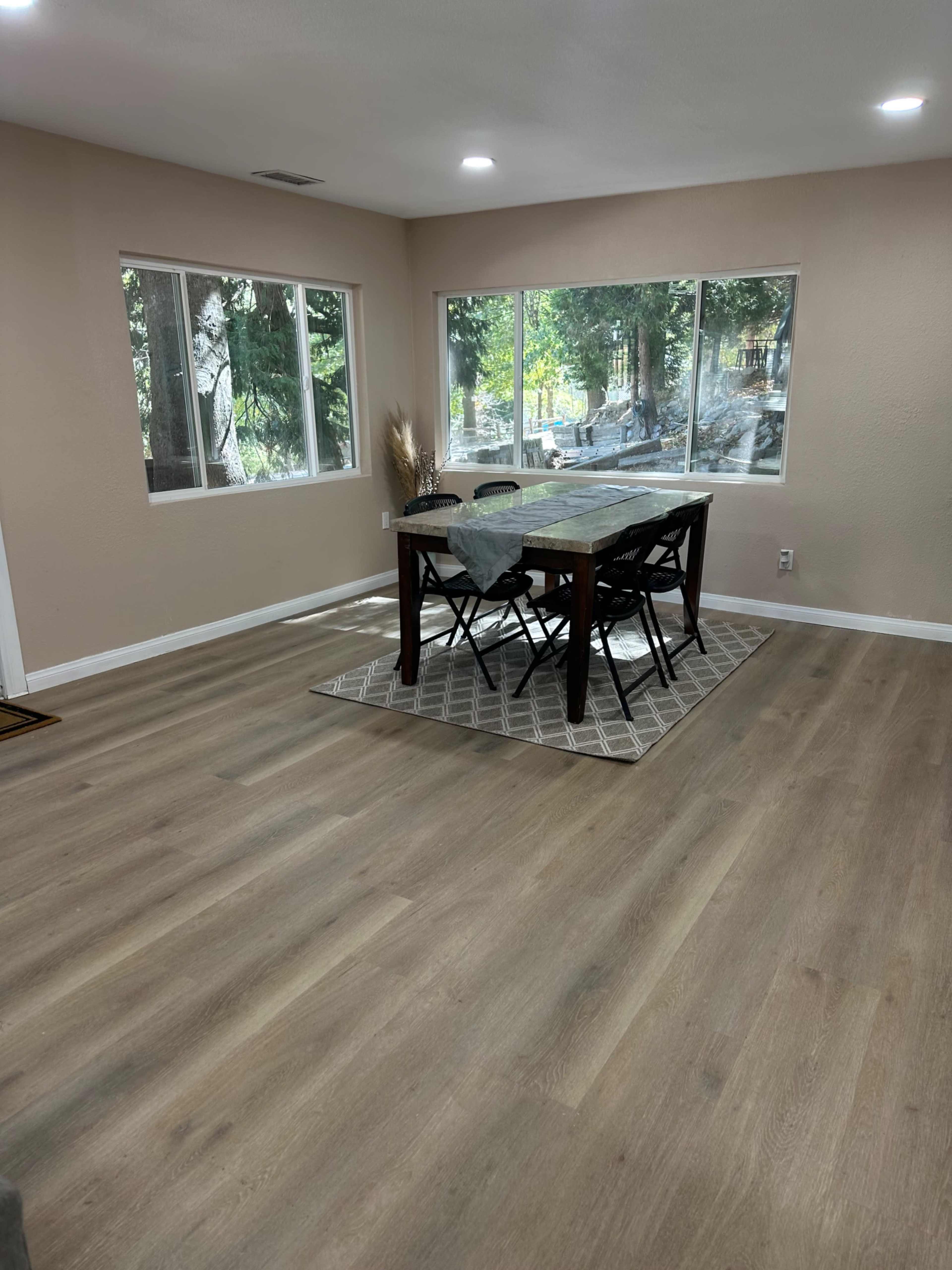 A dining area features a wooden table surrounded by black chairs, set on a patterned rug, with large windows displaying trees outside.