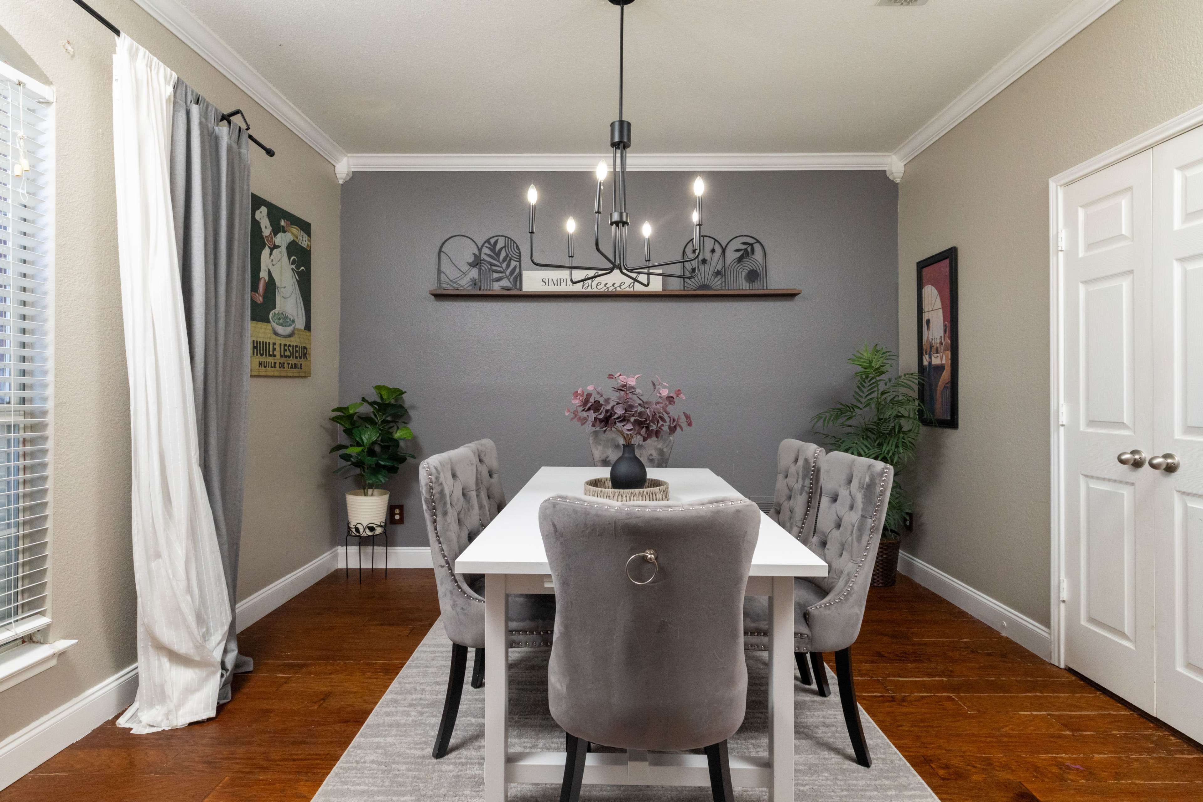 A modern dining room features a white table surrounded by gray upholstered chairs, with decorative wall art and a chandelier above.