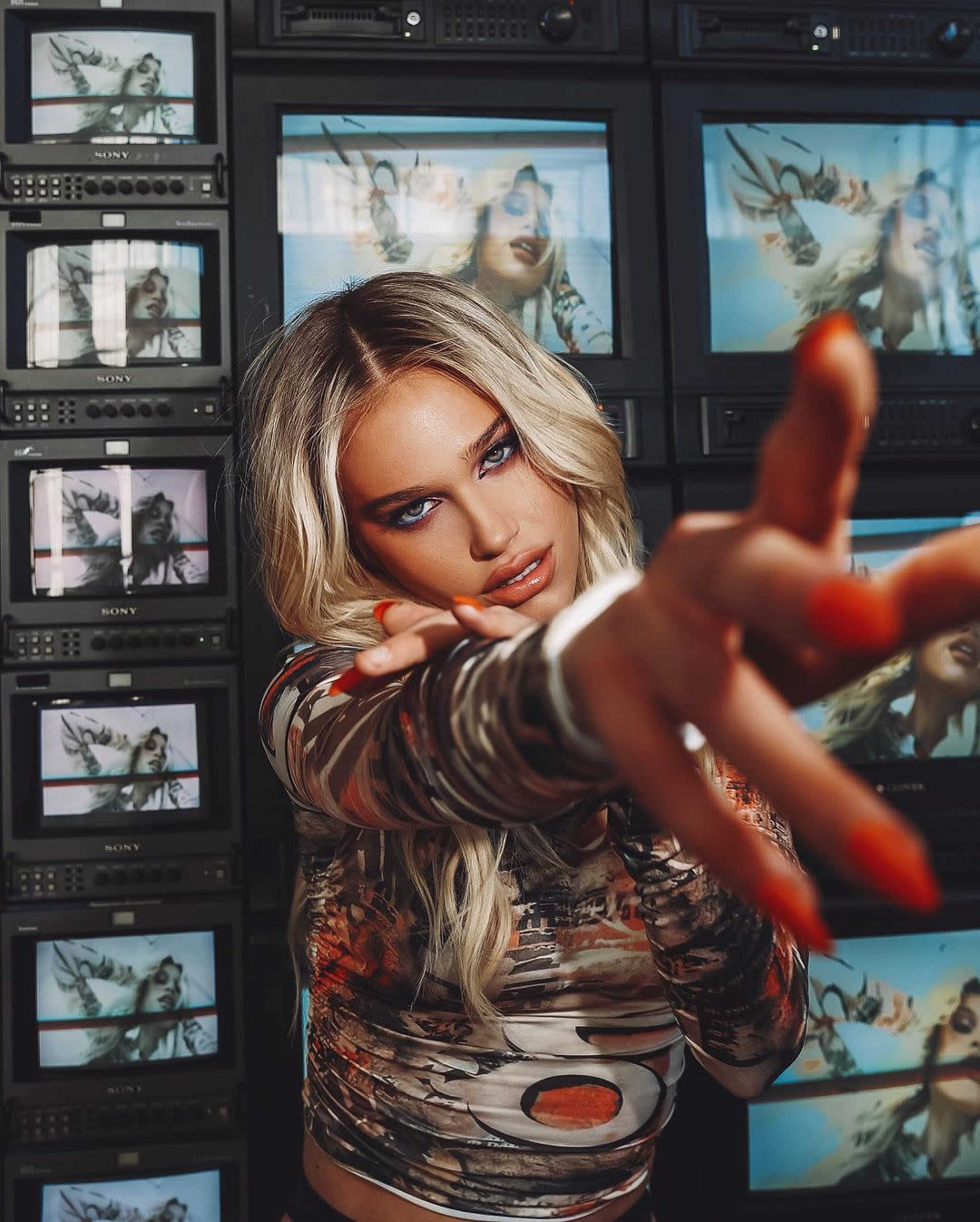 A woman with long, wavy blonde hair extends her arm toward the camera, positioned in front of multiple vintage television screens displaying images of herself.