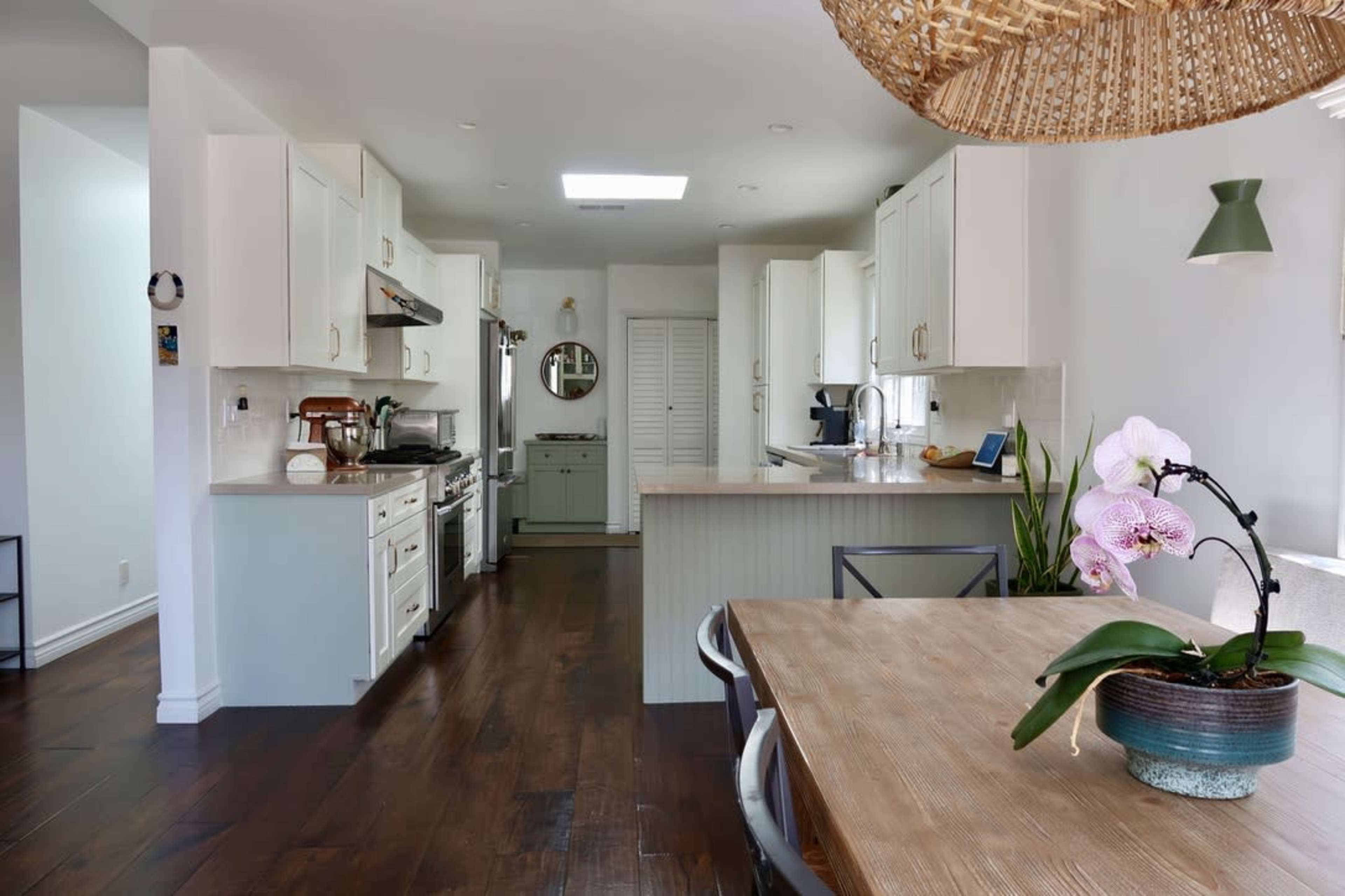 The image shows a modern kitchen with white cabinetry, a wooden dining table, and an orchid on the table, featuring dark wood flooring.