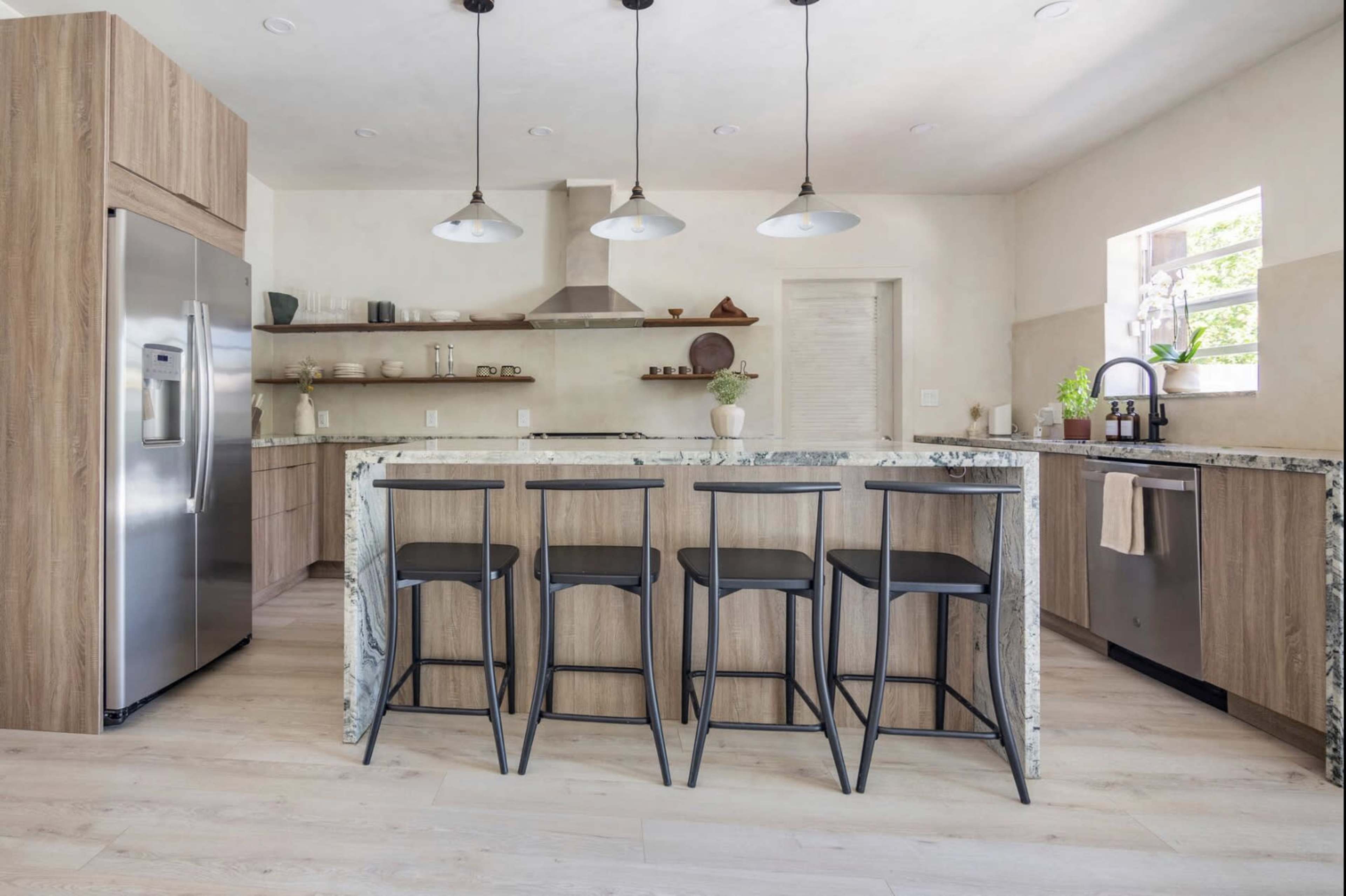 A modern kitchen featuring a large marble-topped island with four black bar stools, stainless steel appliances, and minimalist cabinetry.