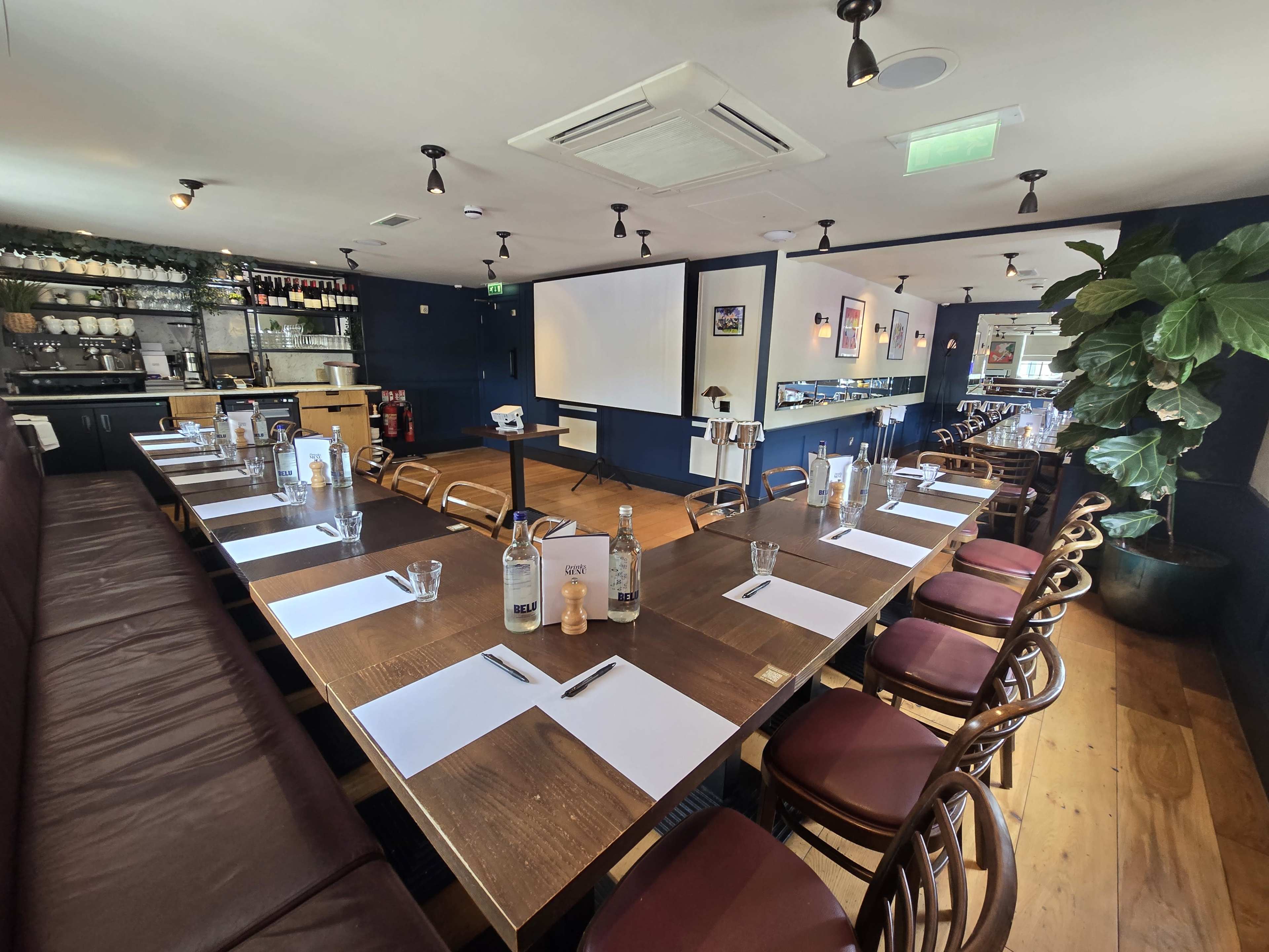The image shows a spacious restaurant interior with a long wooden table set for a meeting, surrounded by brown chairs and decorated with plants and a bar in the background.