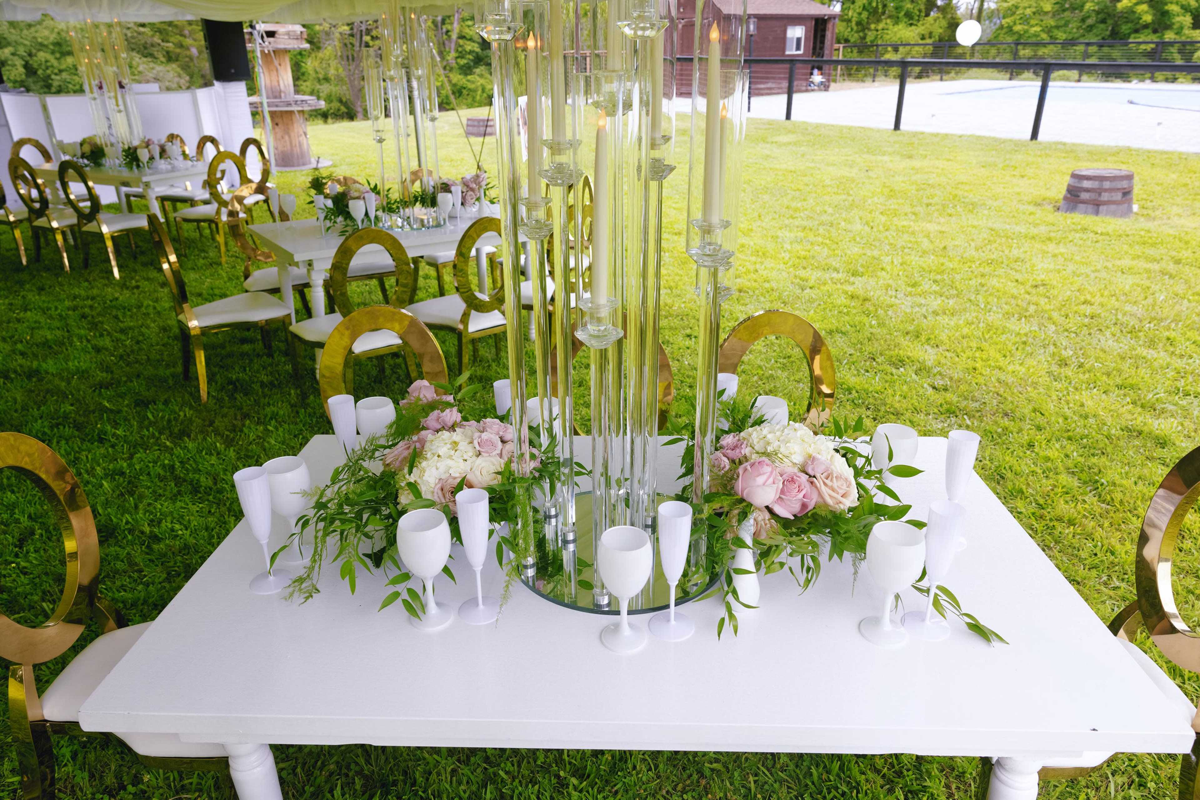 A white dining table adorned with floral arrangements and glass centerpieces is set under a tent on a grassy outdoor venue.