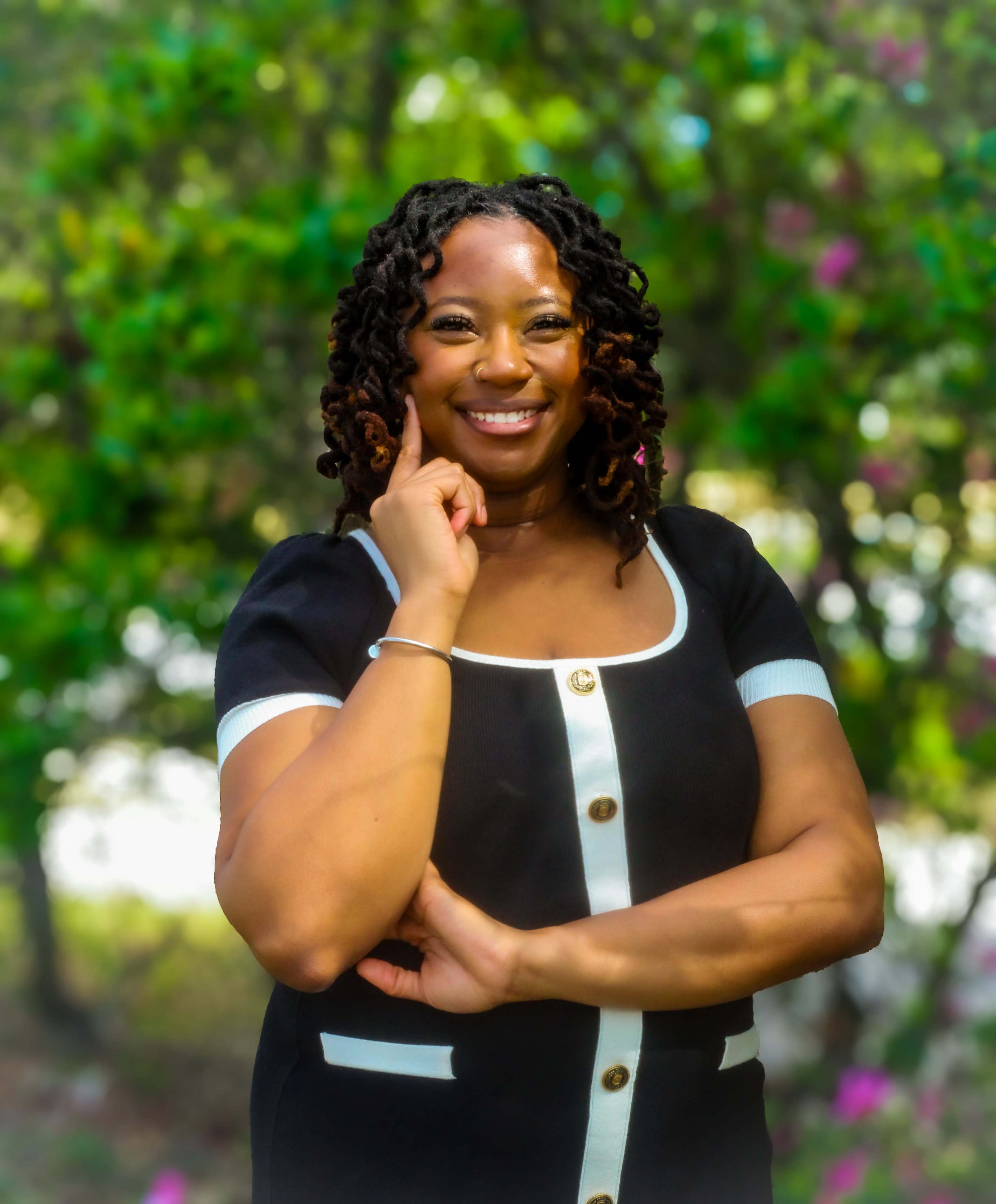 A woman with curly hair is smiling and posing confidently in a black dress with white details against a backdrop of greenery and blooming flowers.