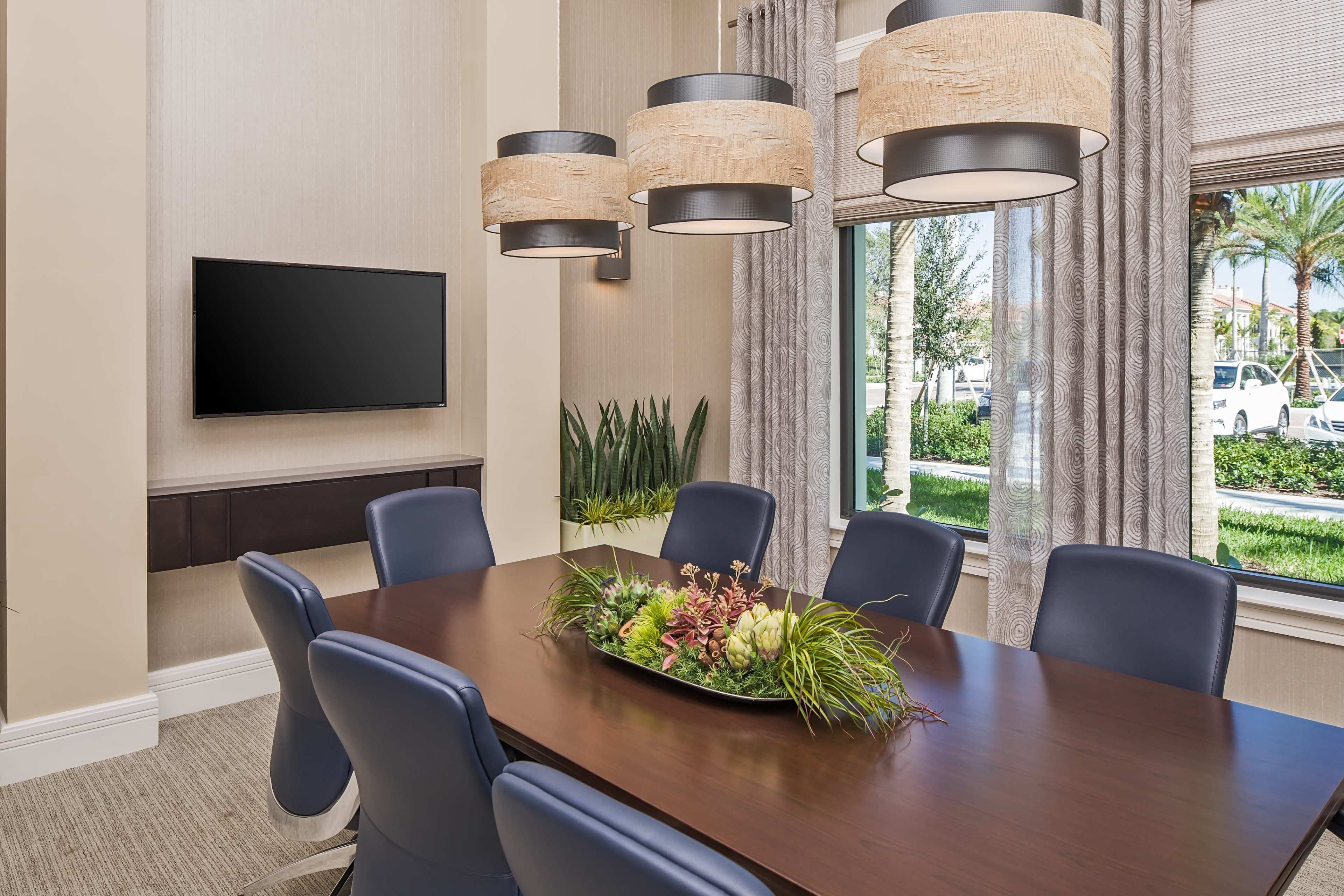 A modern conference room features a long wooden table surrounded by blue chairs, with a TV mounted on the wall and large windows displaying greenery outside.