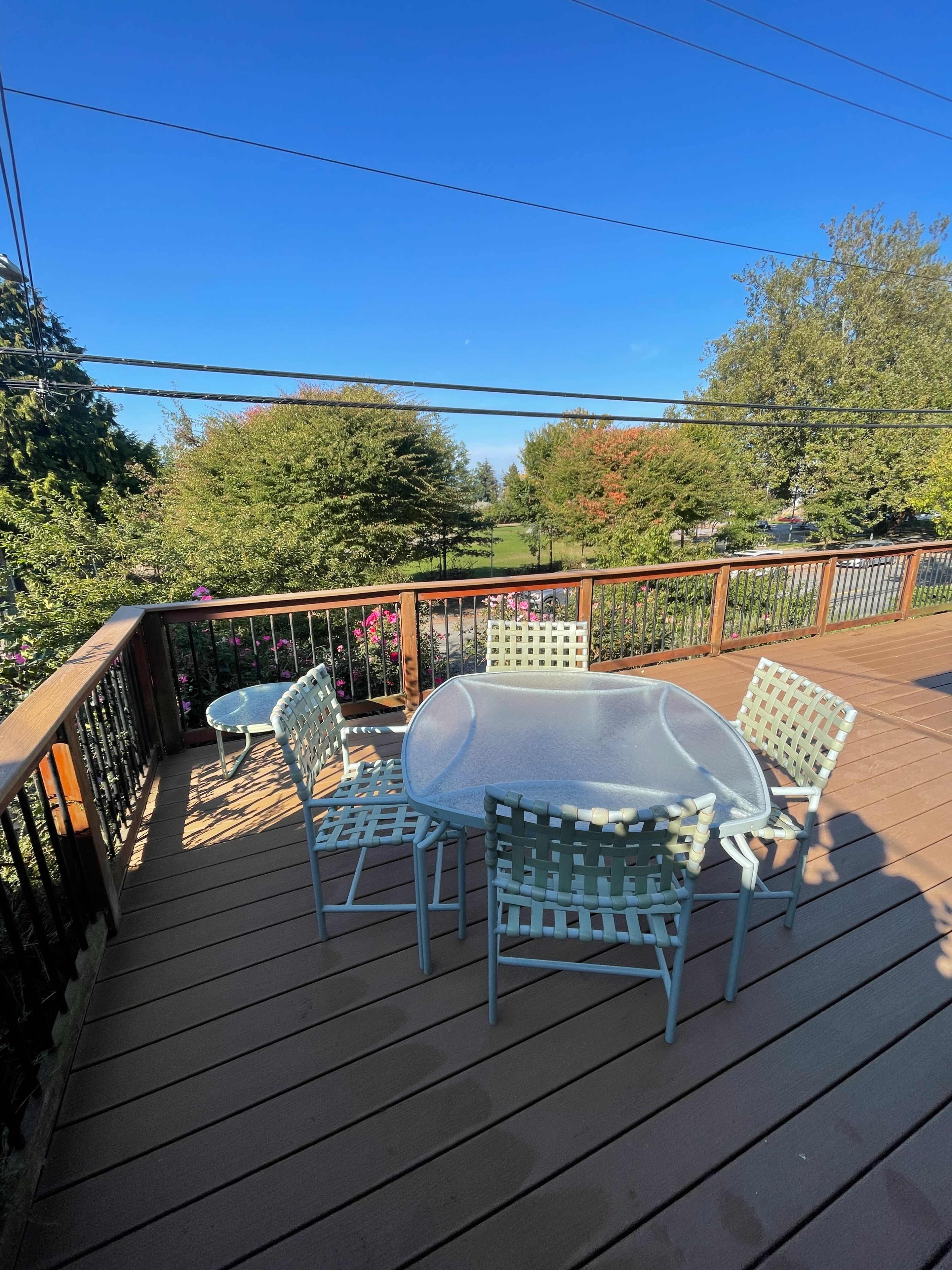 A wooden deck features a glass-top table surrounded by six light-colored chairs, with trees and a clear sky visible in the background.