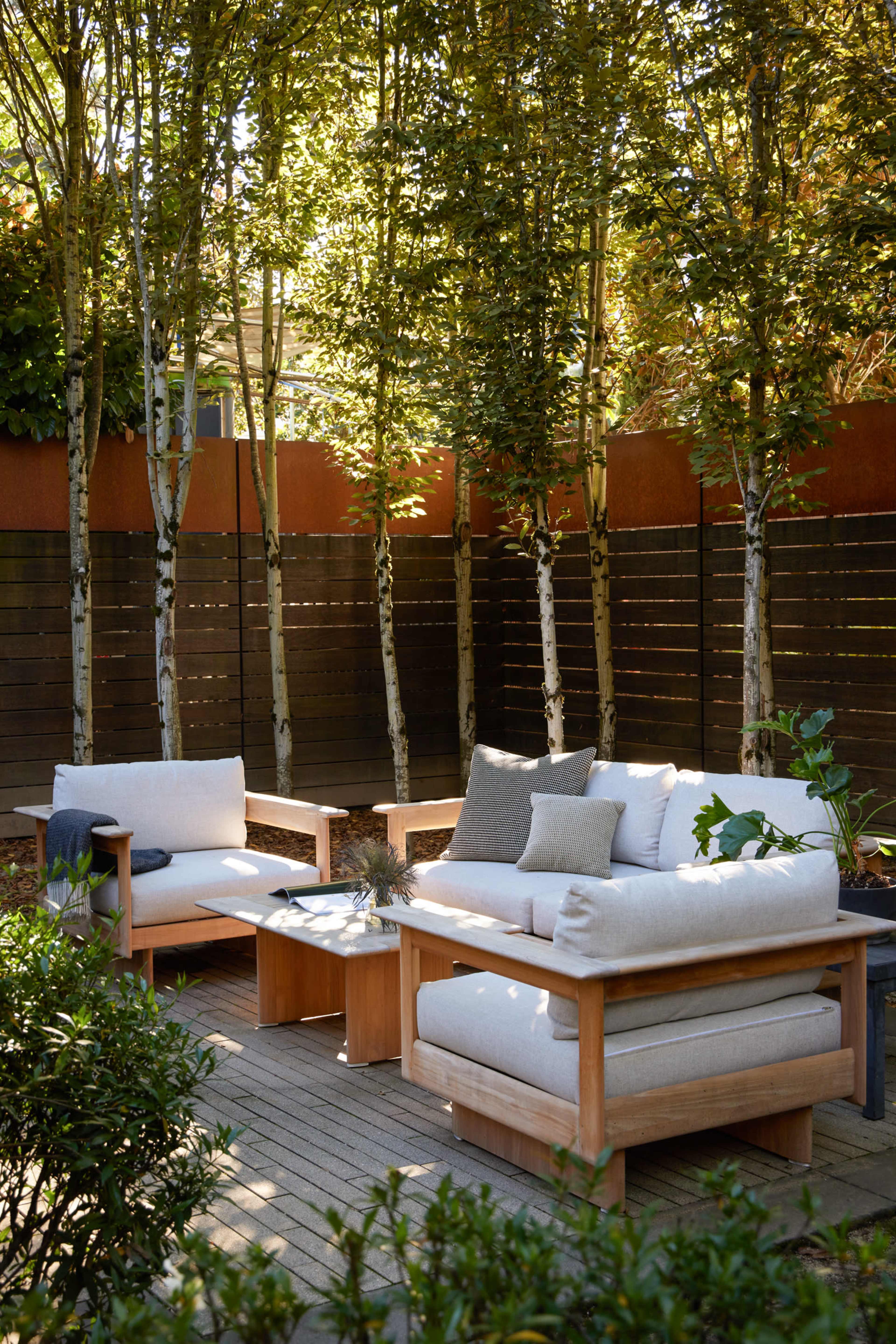 A cozy outdoor seating area features wooden furniture with soft cushions surrounded by tall trees and a wooden fence.