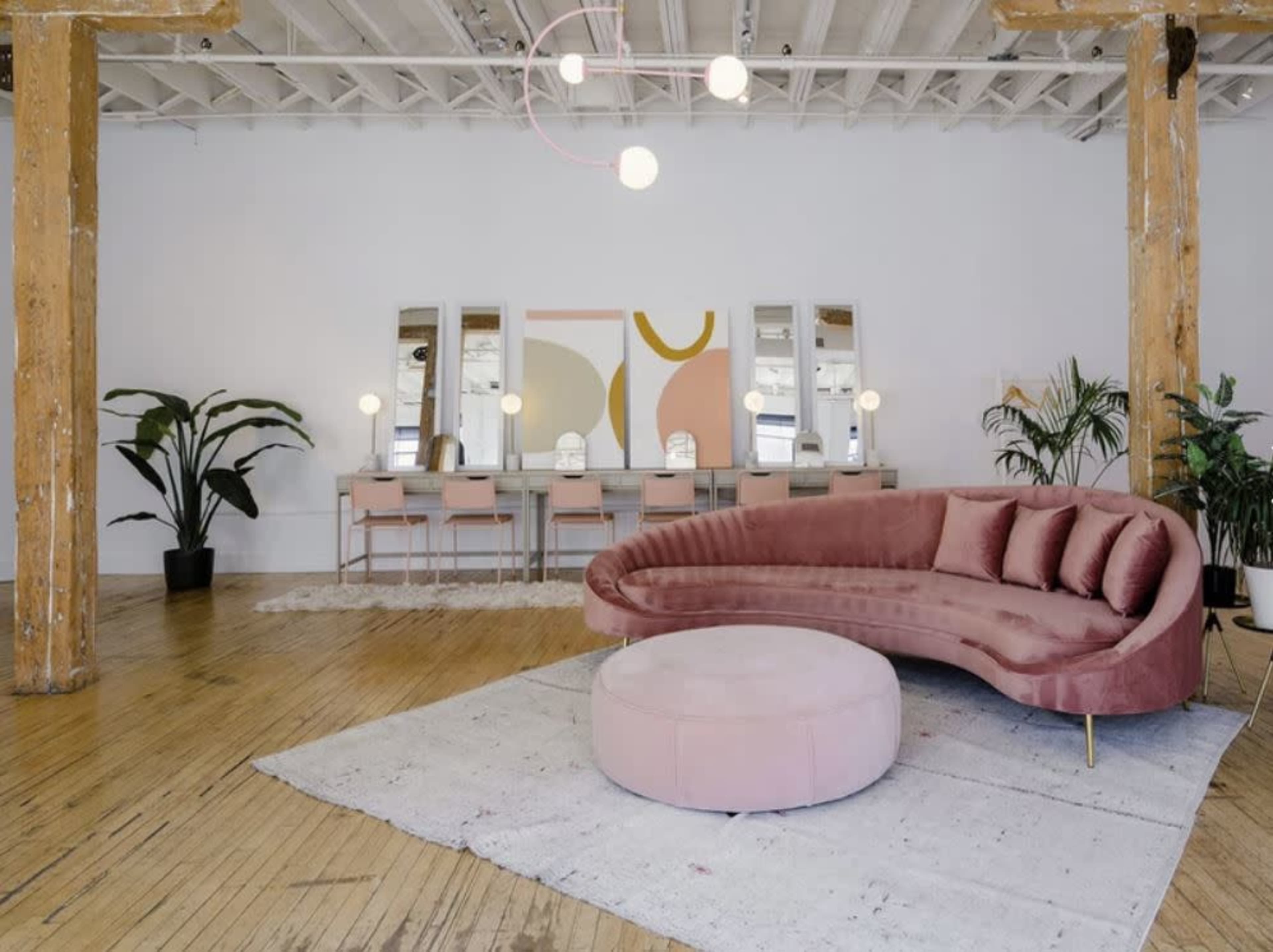 A modern interior room with a curved pink sofa, a round ottoman, a long table with chairs, and large mirrors against a white wall.