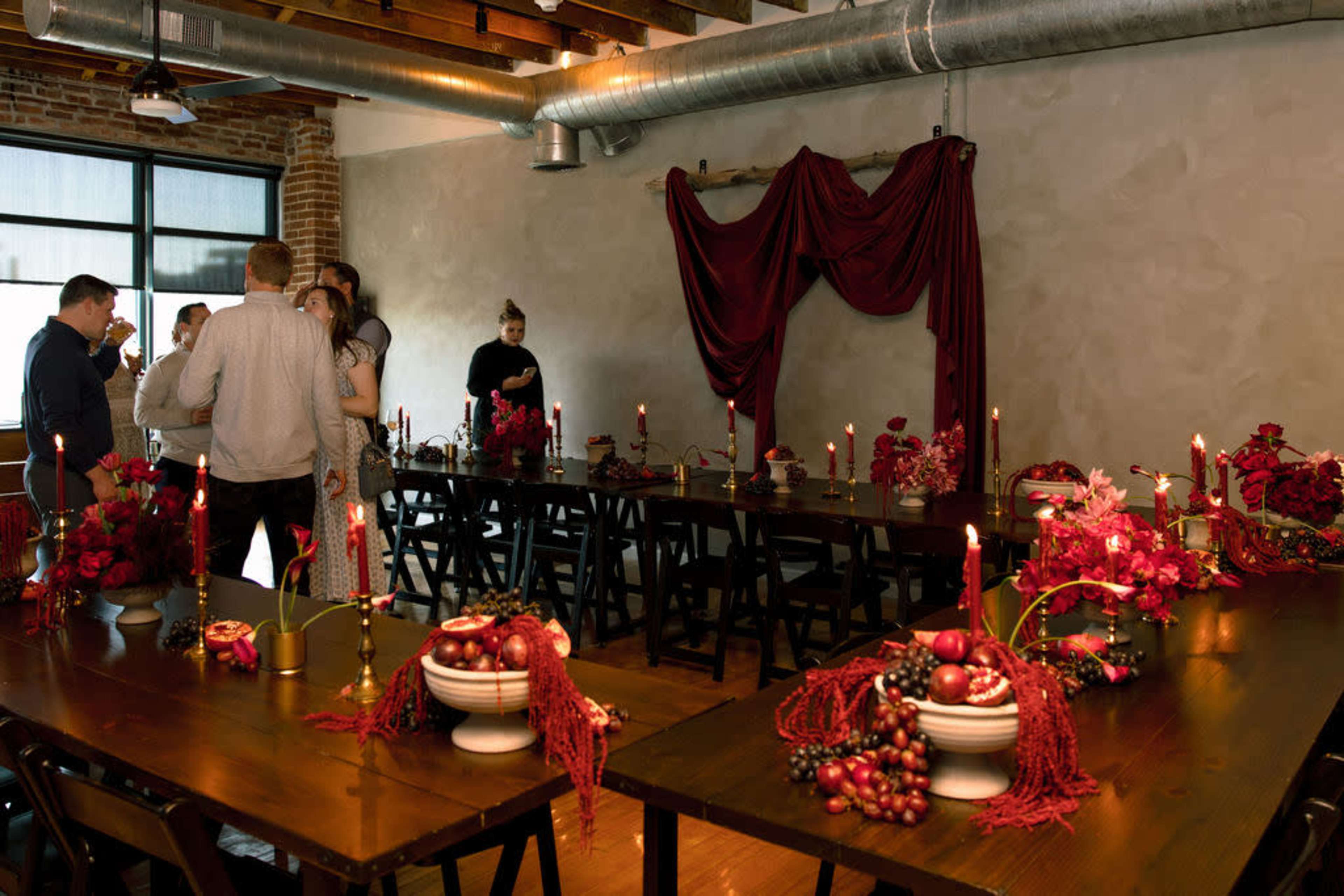 The image shows a dining area with a long table set for an event, decorated with red floral arrangements and lit candles, while people mingle in the background.