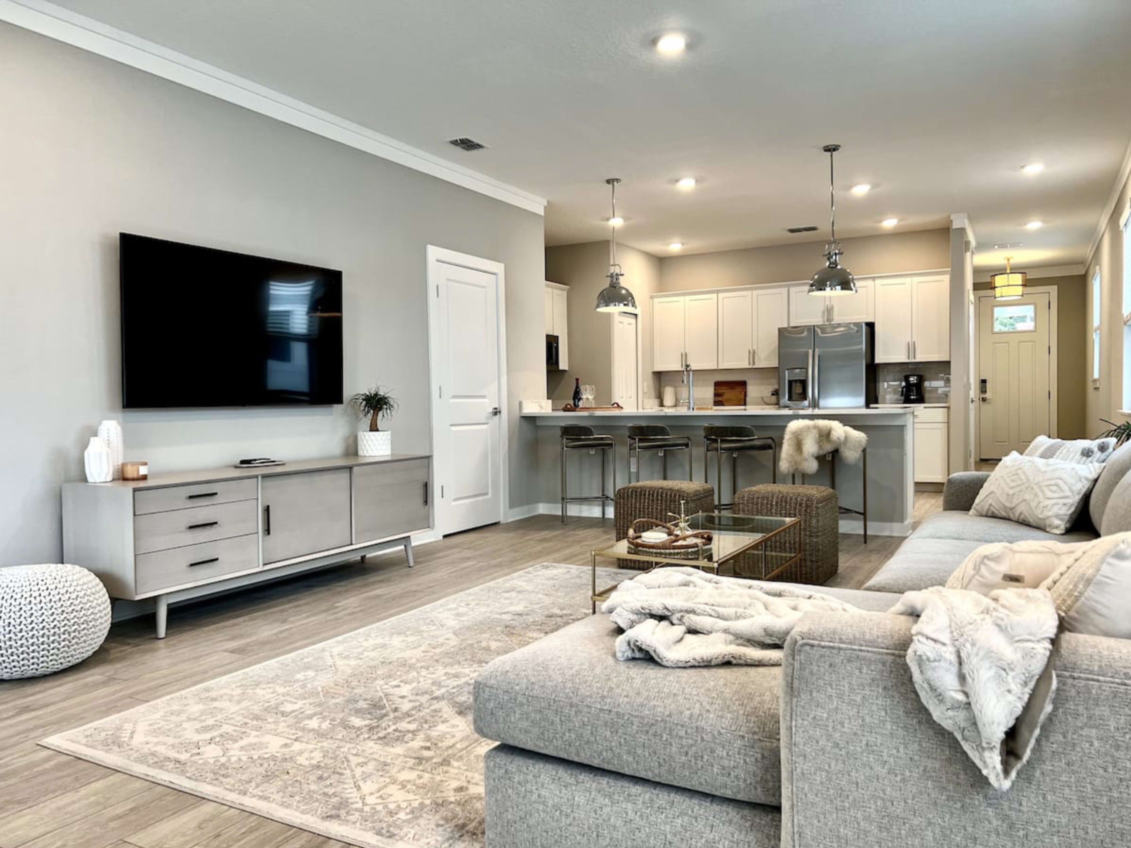 A modern living room with a light gray sofa, a flat-screen TV mounted on the wall, and a kitchen area featuring white cabinets and bar stools.