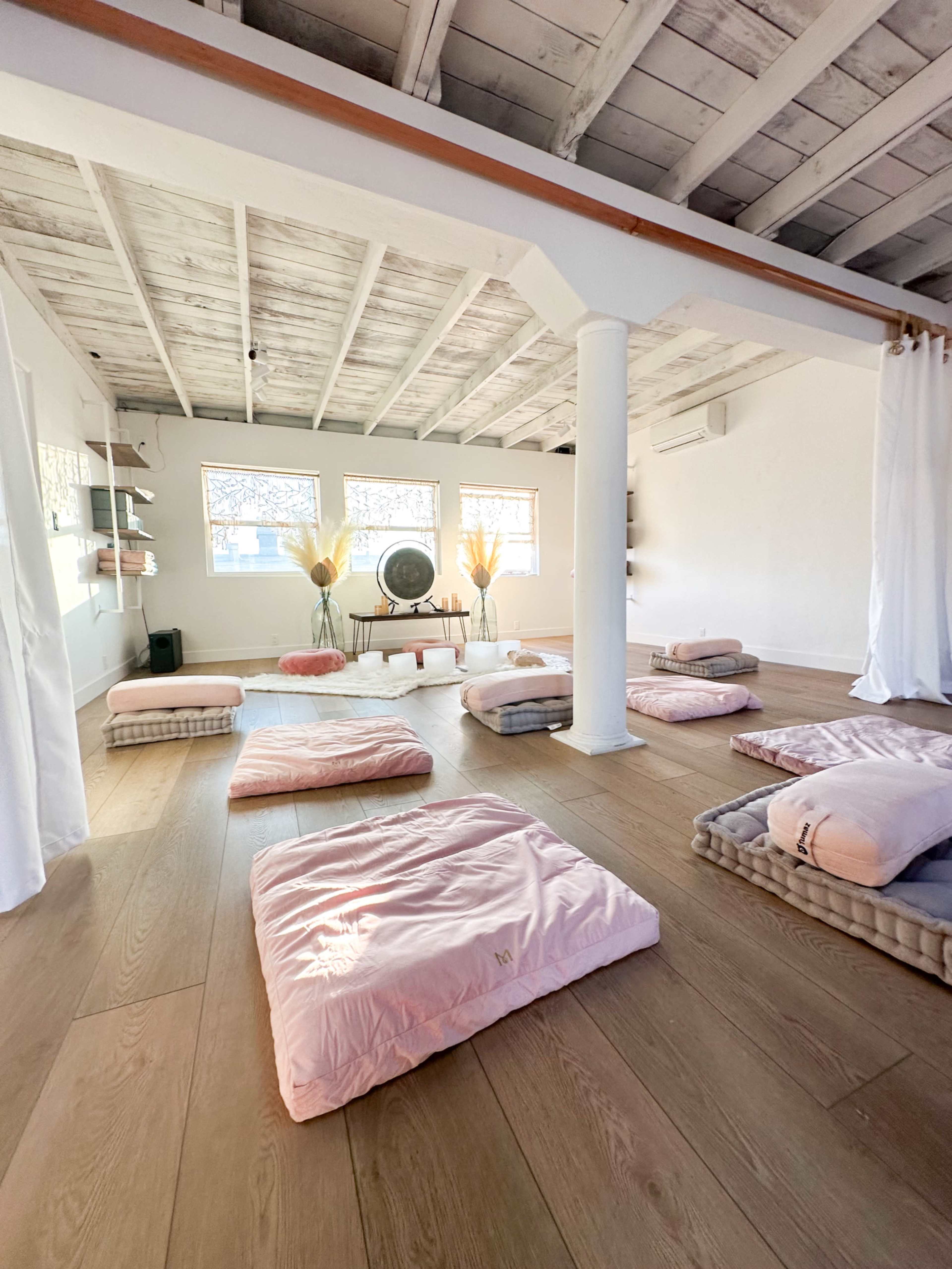 A spacious, minimalist meditation room with several pink mats arranged on a wooden floor, complemented by soft lighting and a large wall fan.