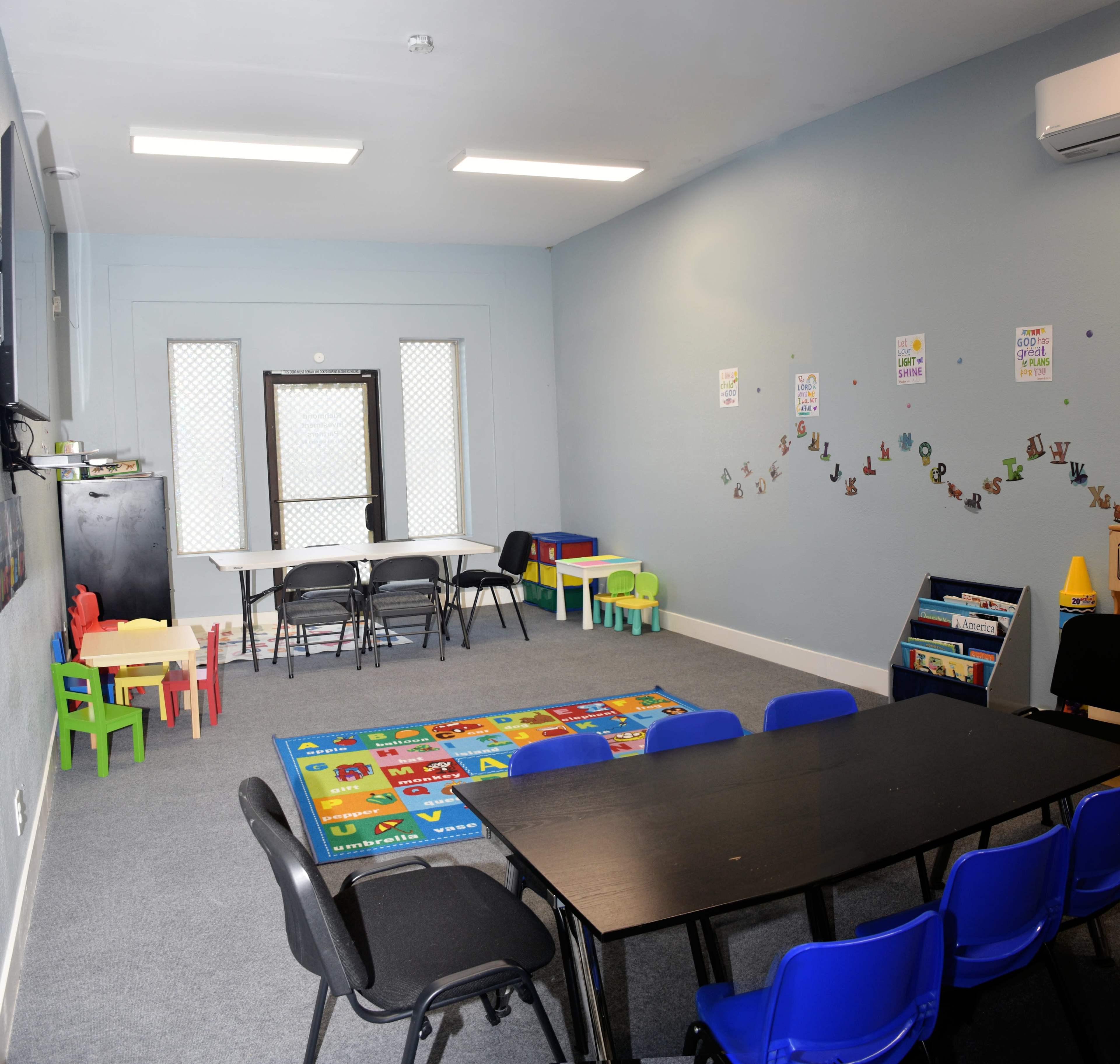 A classroom is shown with a large rug, a black table with blue chairs, smaller tables with chairs, and educational posters on the walls.
