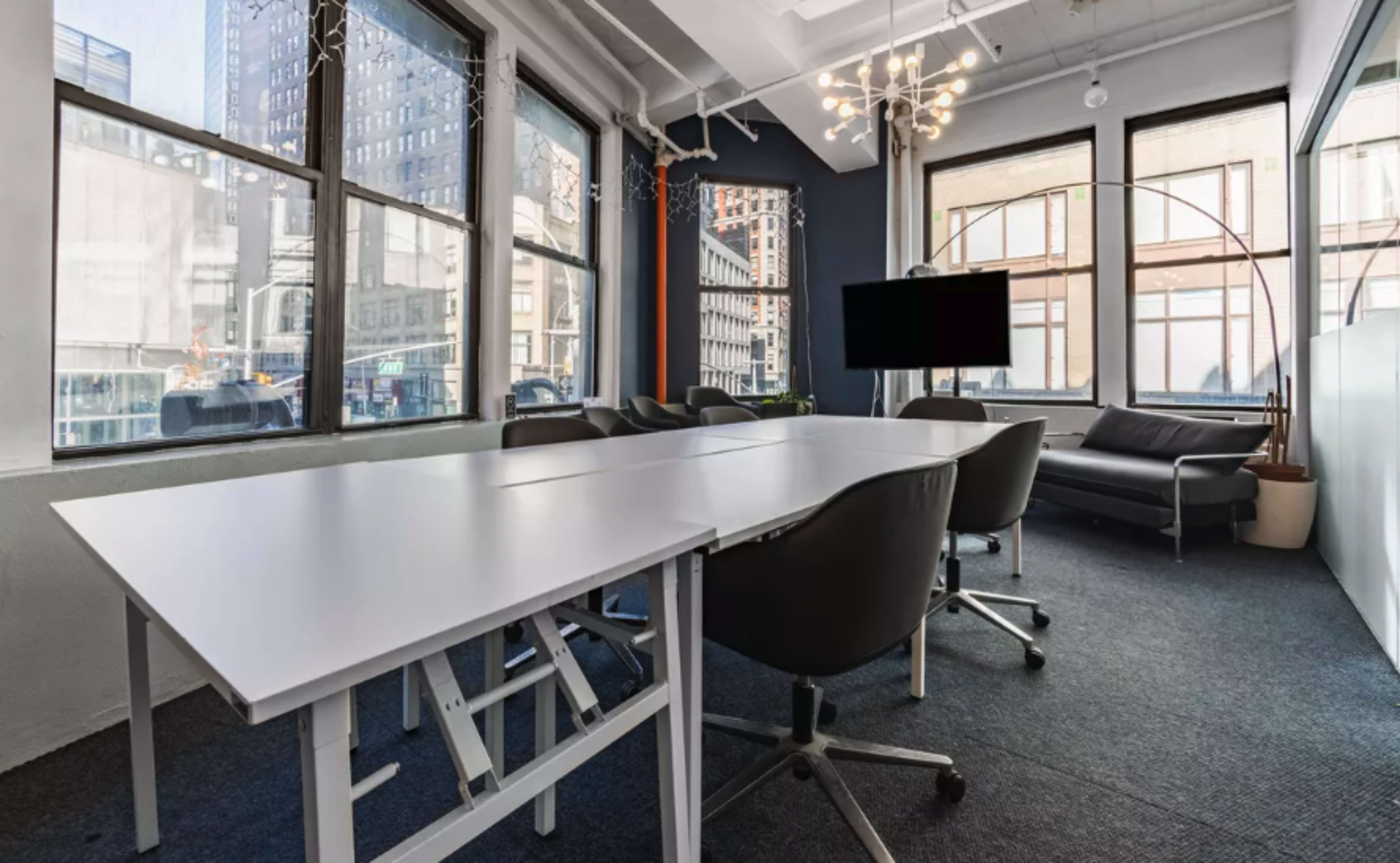 A modern conference room features a white table surrounded by ergonomic chairs, large windows revealing an urban skyline, and a wall-mounted television.