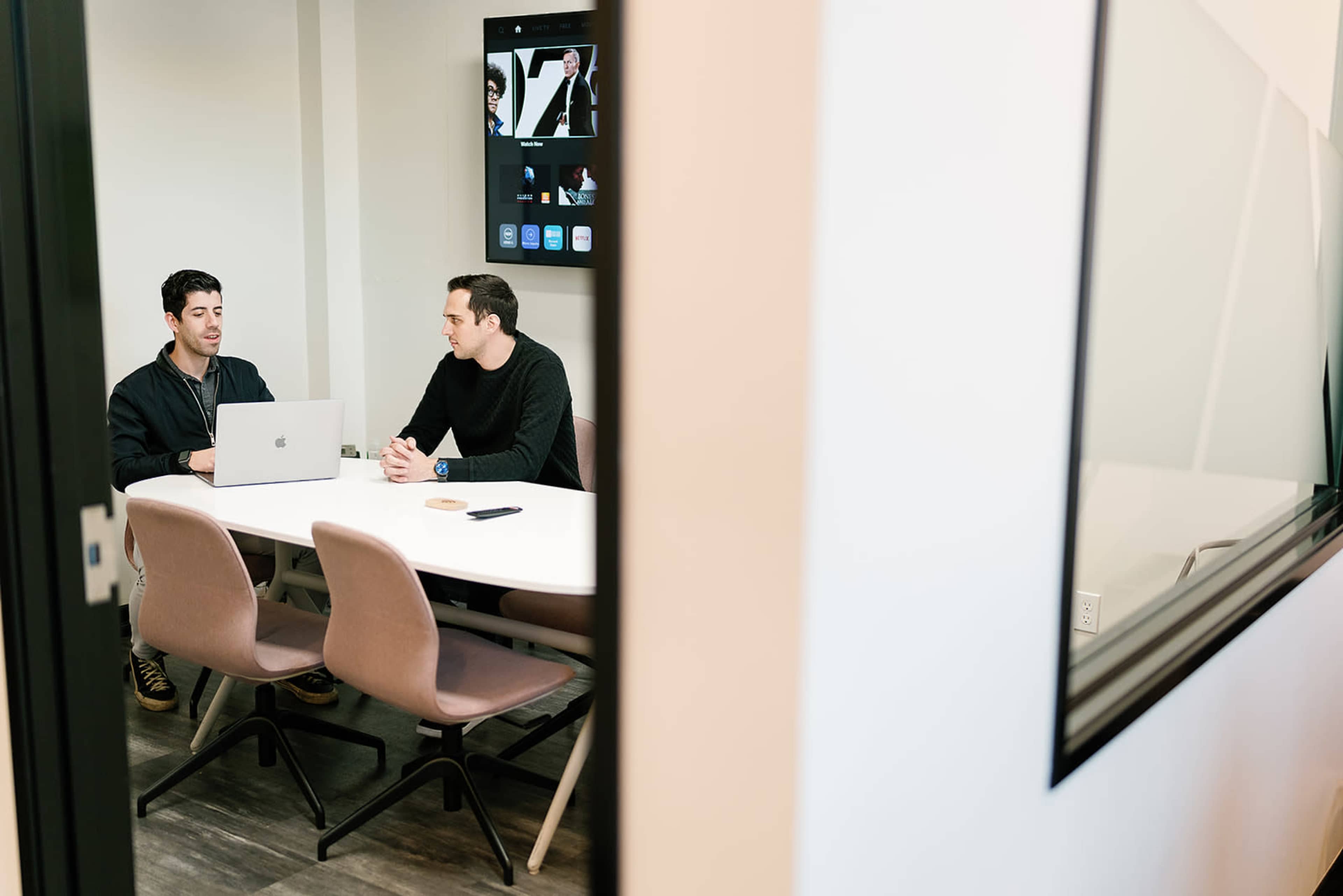 Two men are seated at a conference table with a laptop between them in a modern meeting room.