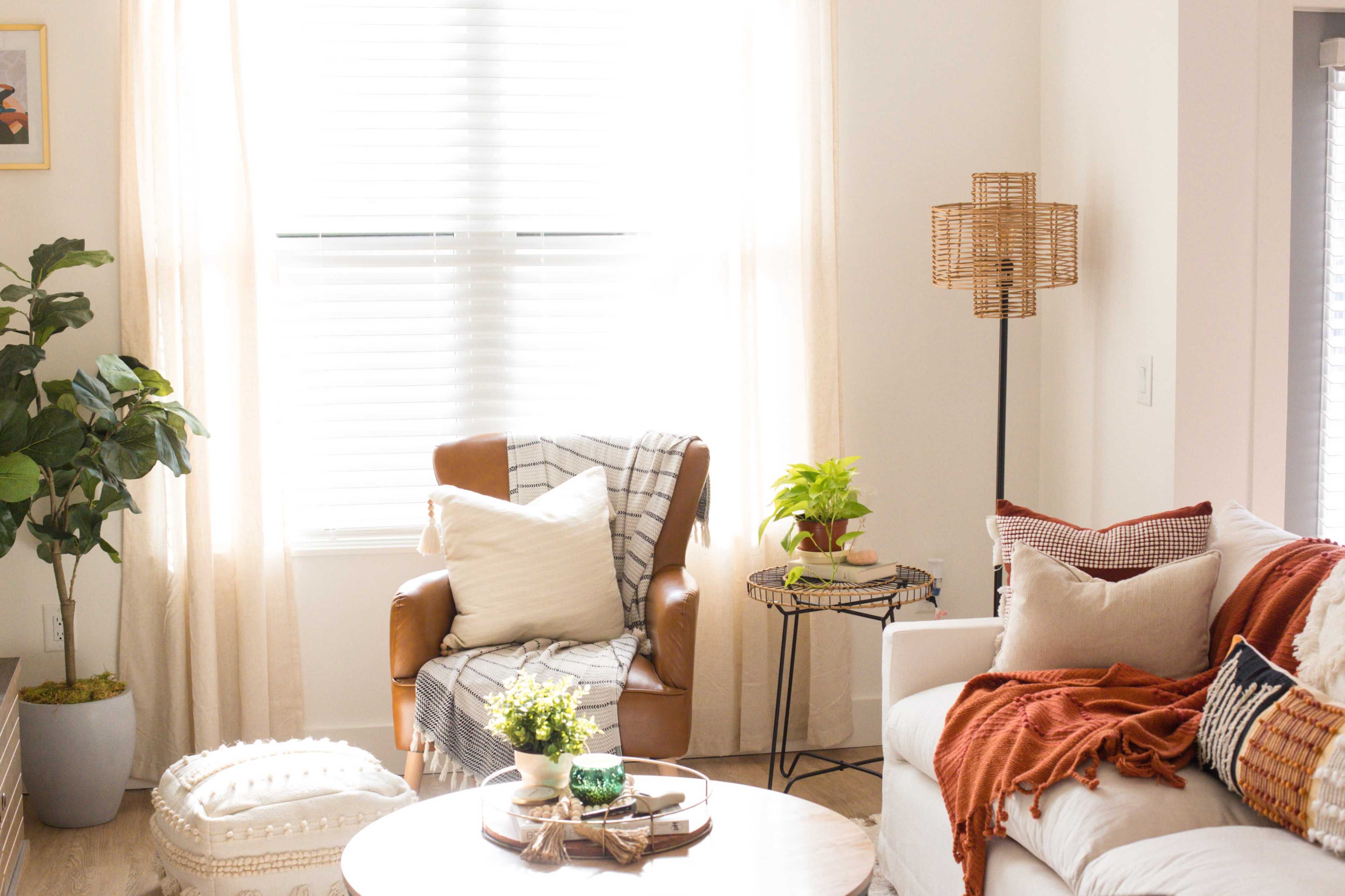A cozy living room features a brown armchair with a plaid throw, a white sofa adorned with decorative pillows, and a coffee table surrounded by houseplants and natural light.
