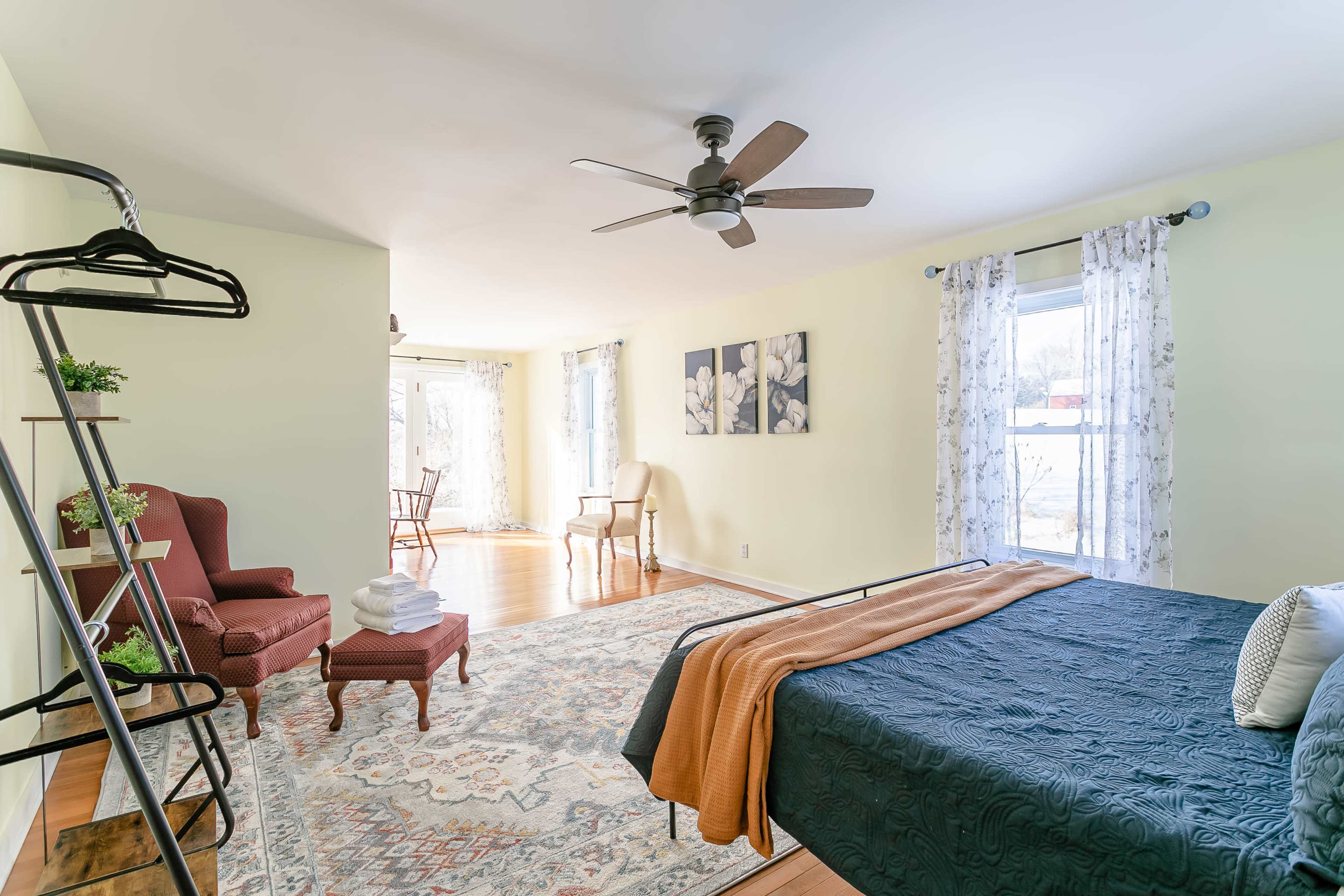 A bright bedroom featuring a bed with a patterned blanket, a seated area with a chair, a small table, and a decorative rug on wooden flooring.