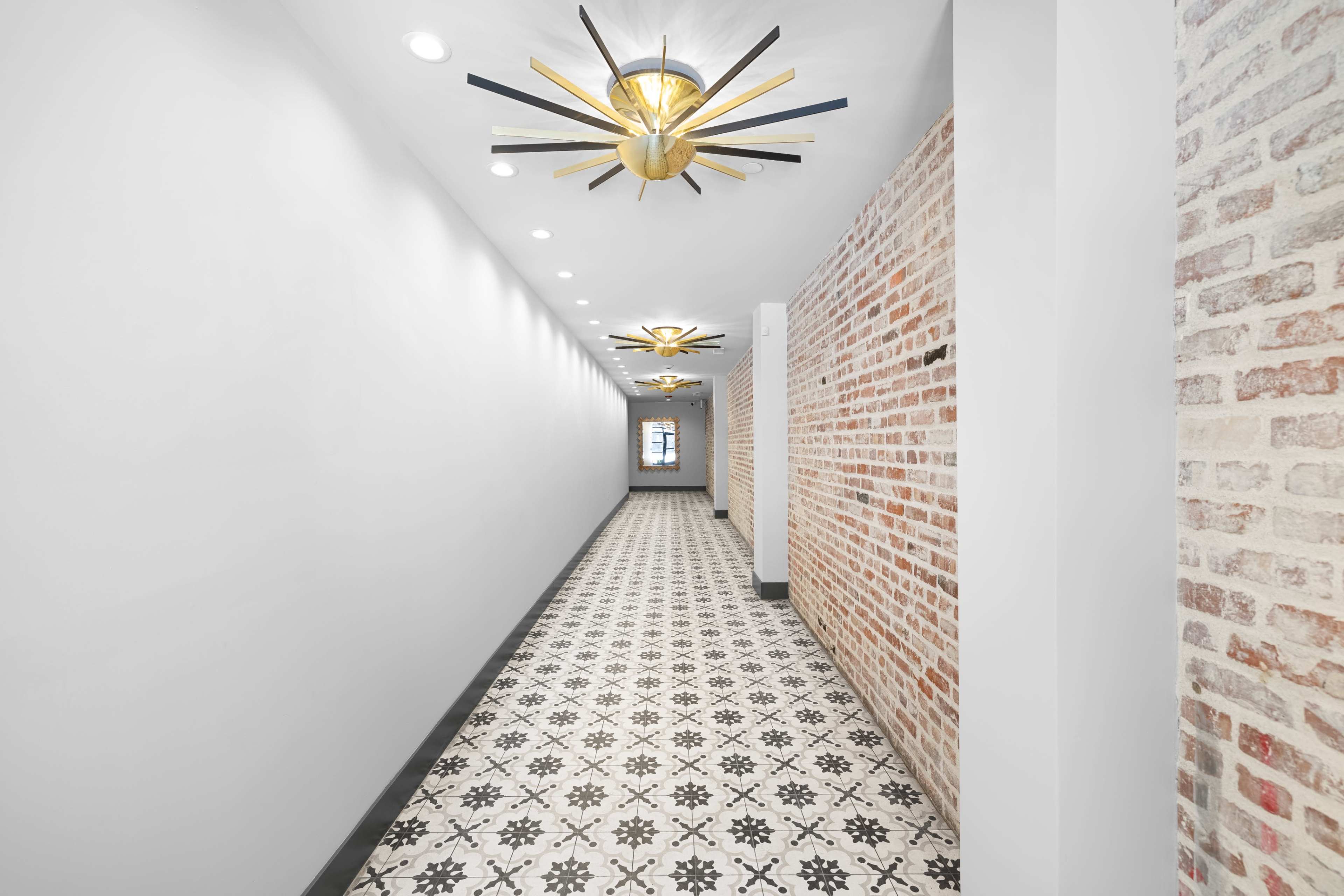 The image shows a long, narrow hallway with a patterned tile floor, exposed brick walls, and modern light fixtures overhead.