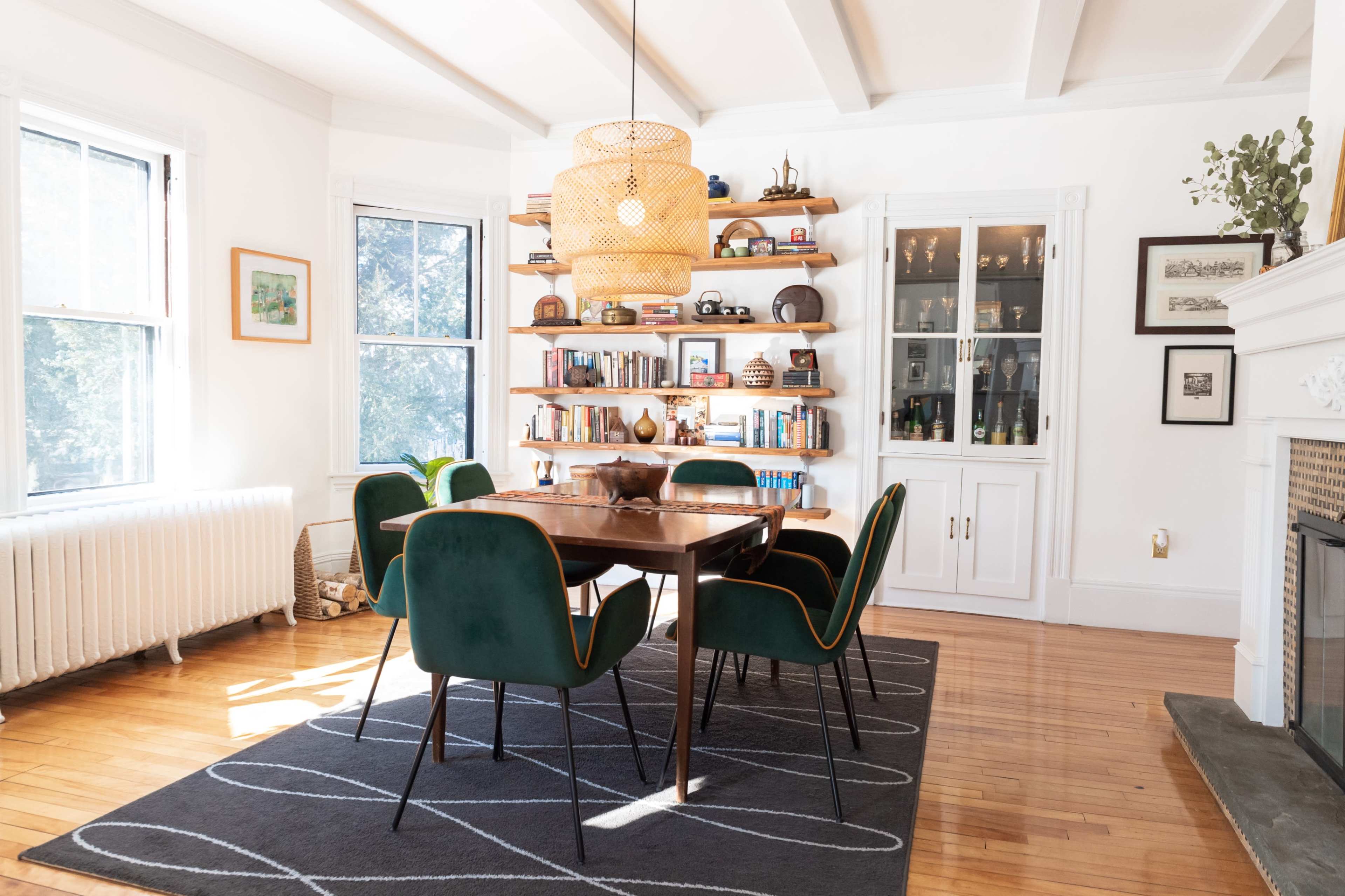 A dining room features a wooden table surrounded by green chairs, with bookshelves and a large light fixture overhead.