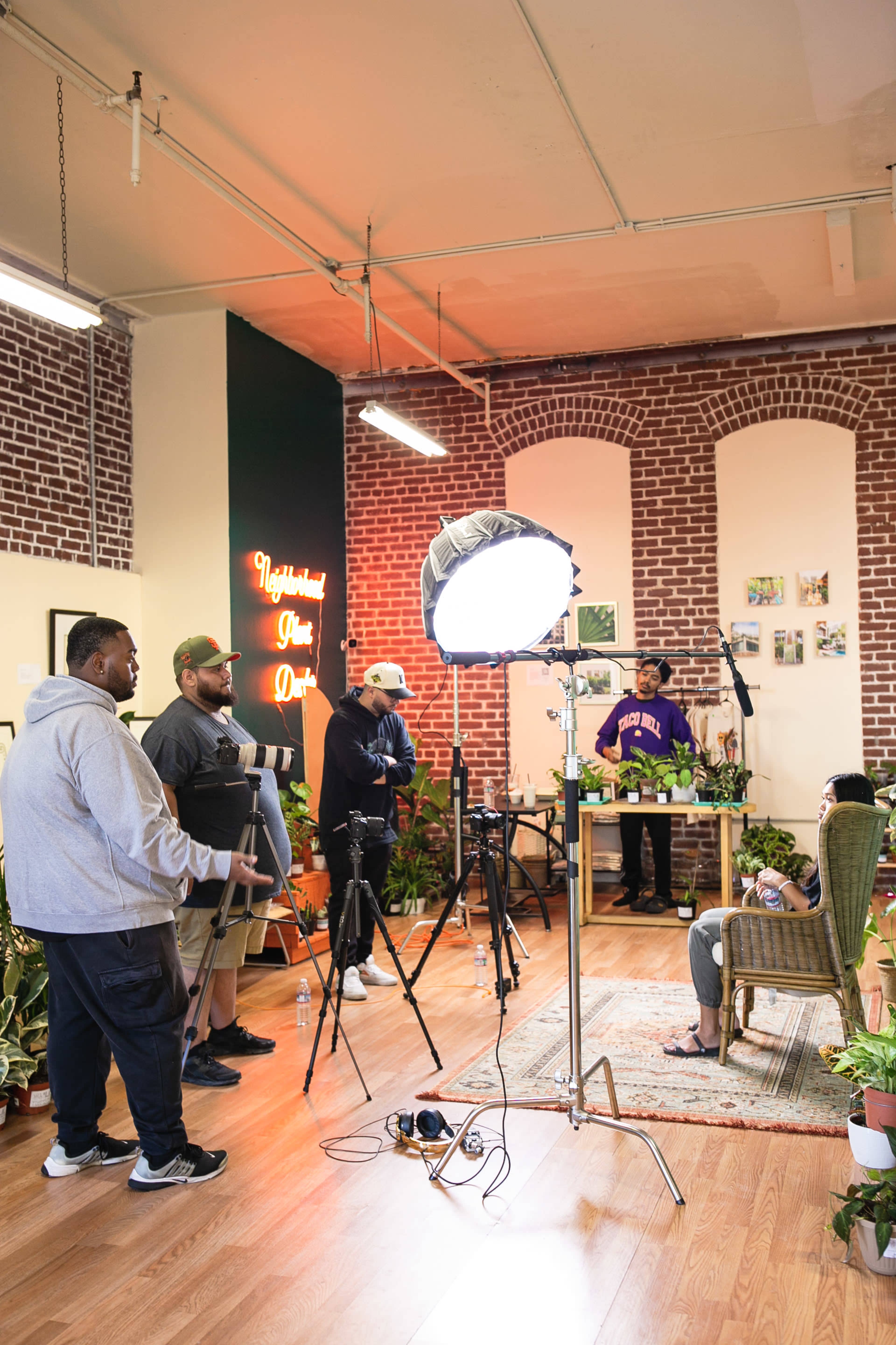 A group of four individuals sets up video equipment in a room filled with plants and brick walls.