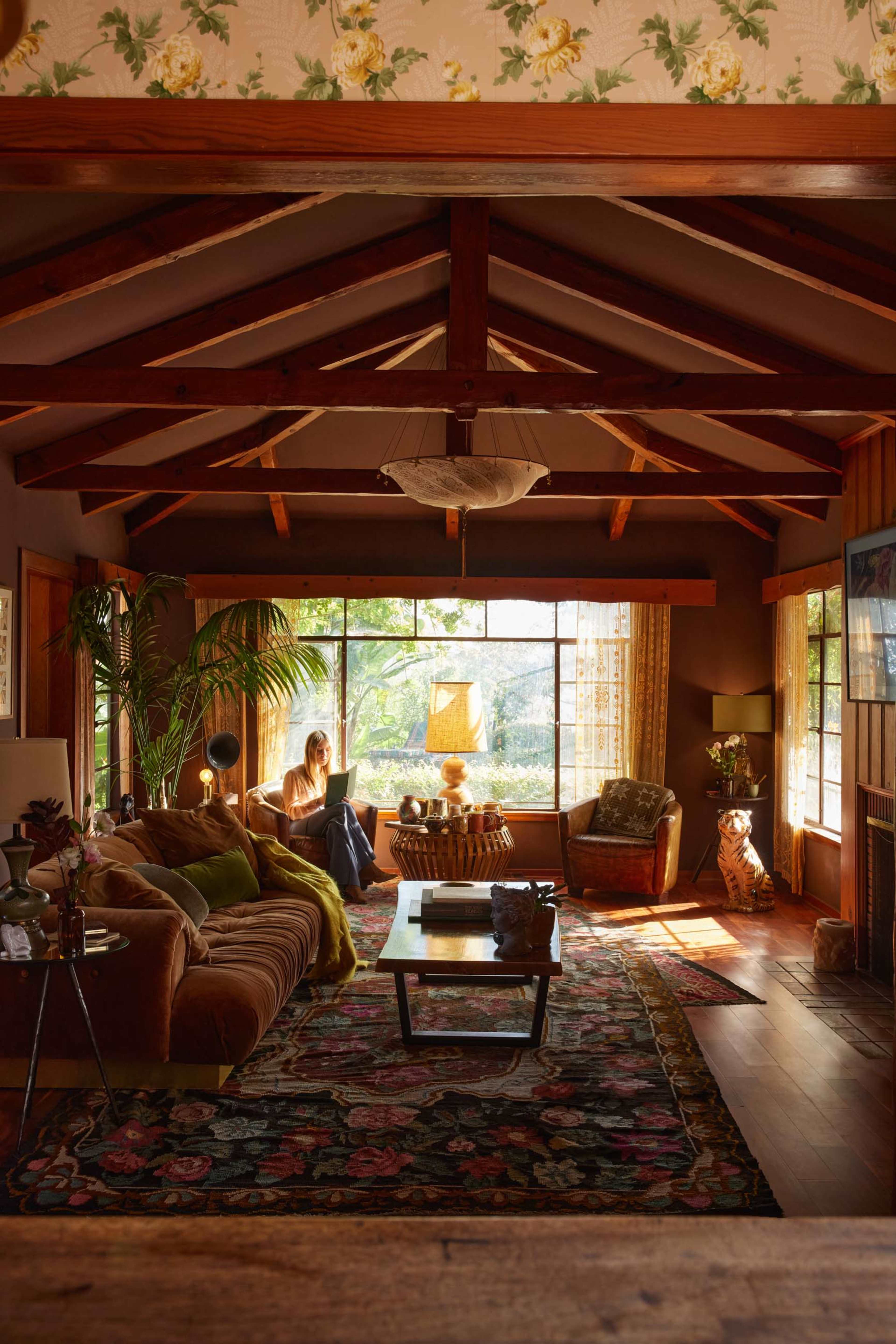 A woman sits on a couch in a sunlit living room with wooden beams, large windows, and a floral rug.