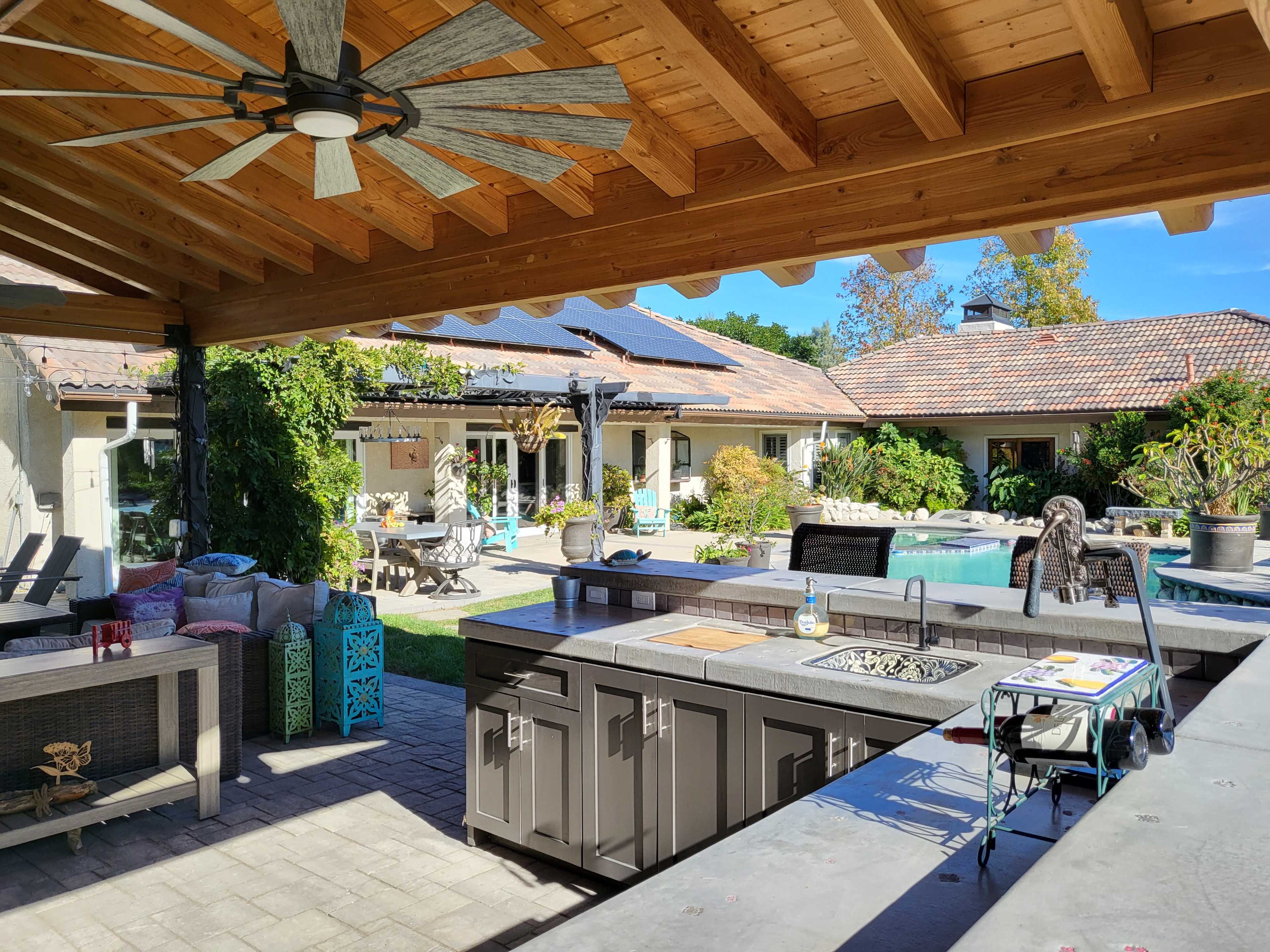 An outdoor kitchen under a wooden roof with a ceiling fan, overlooking a swimming pool and a landscaped backyard area with various seating options.