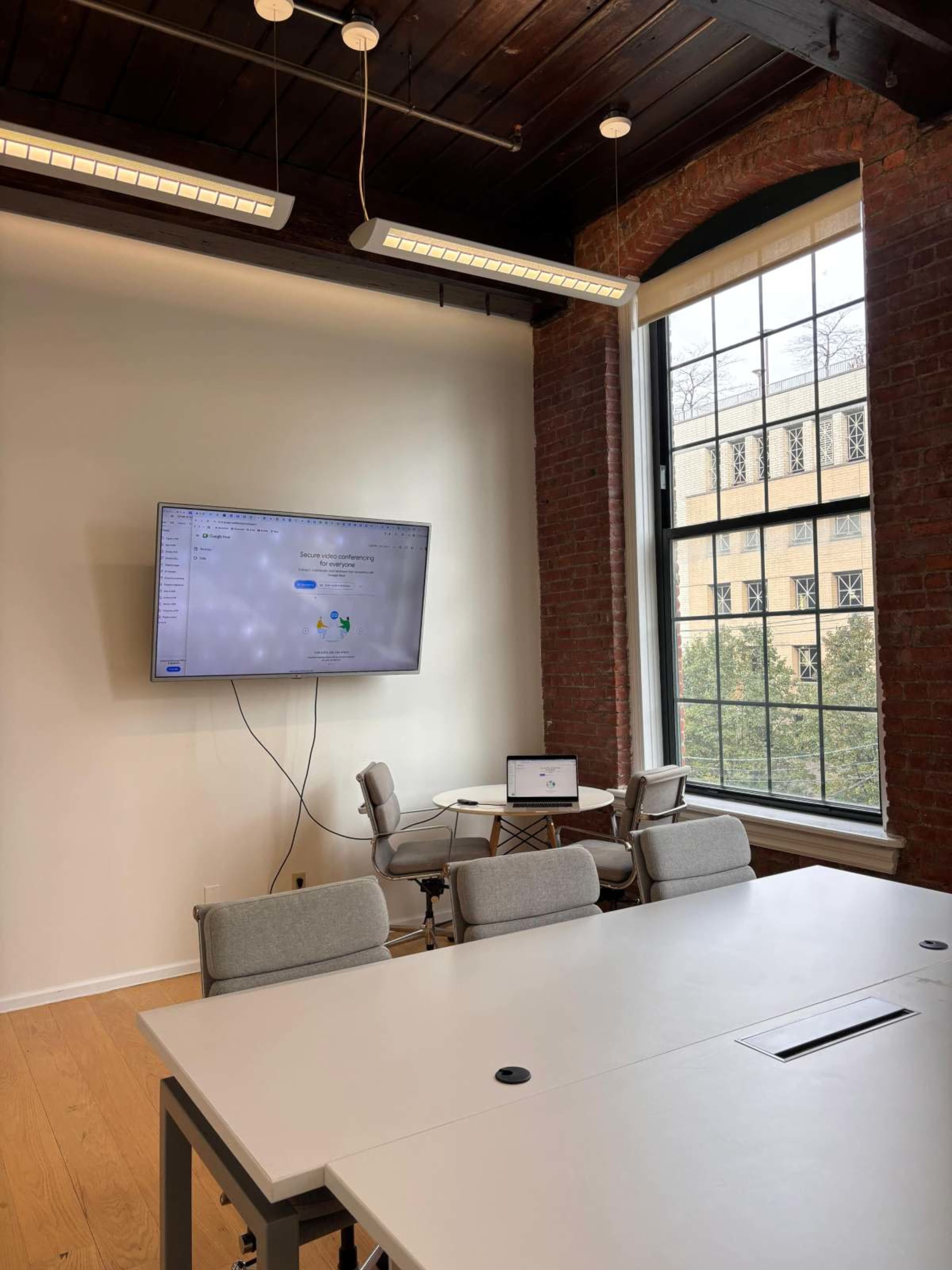 The image shows a modern office conference room with a large screen mounted on the wall, a table with chairs, and a window providing natural light.