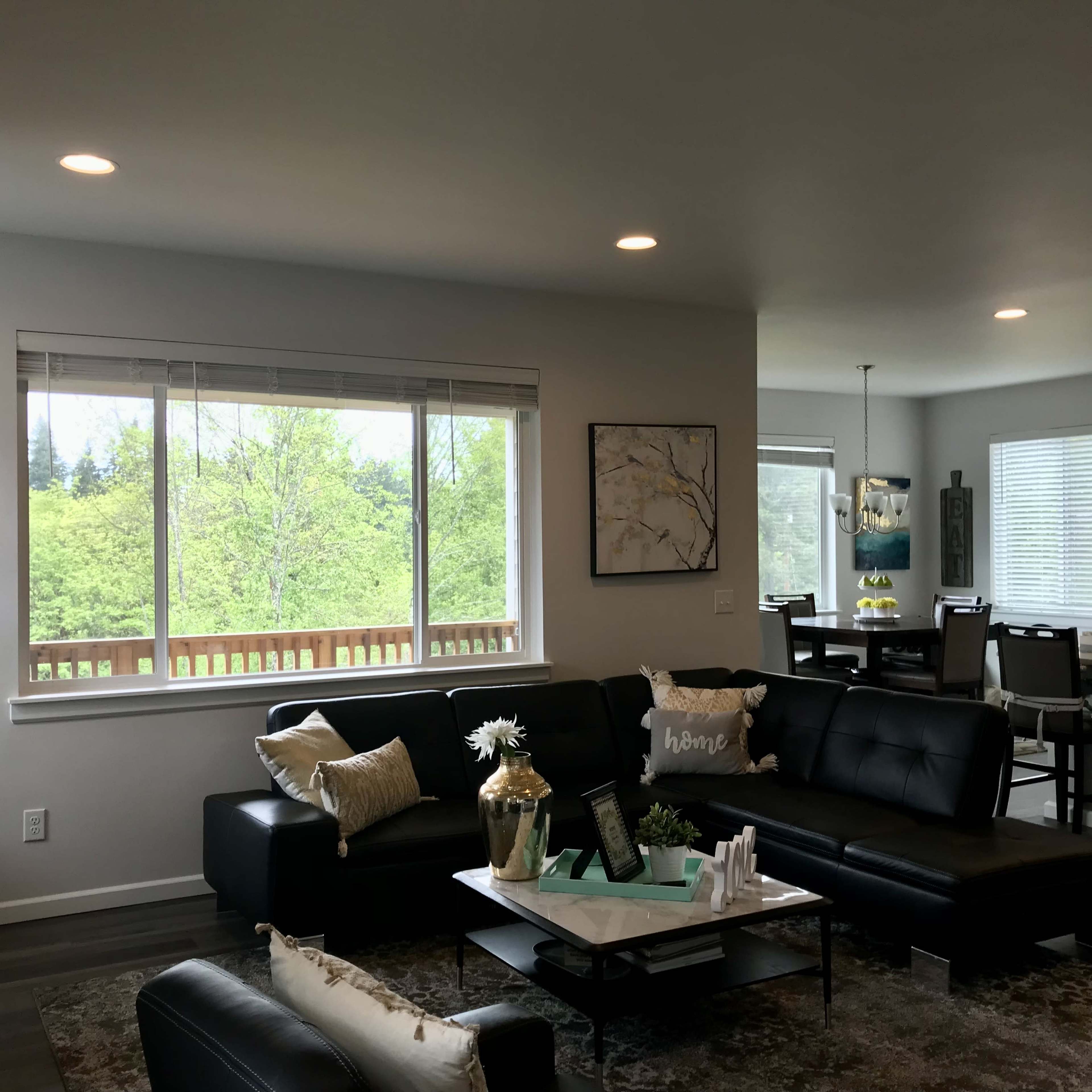 A modern living room features a black sectional sofa, a coffee table with decorative items, and large windows overlooking a green landscape.
