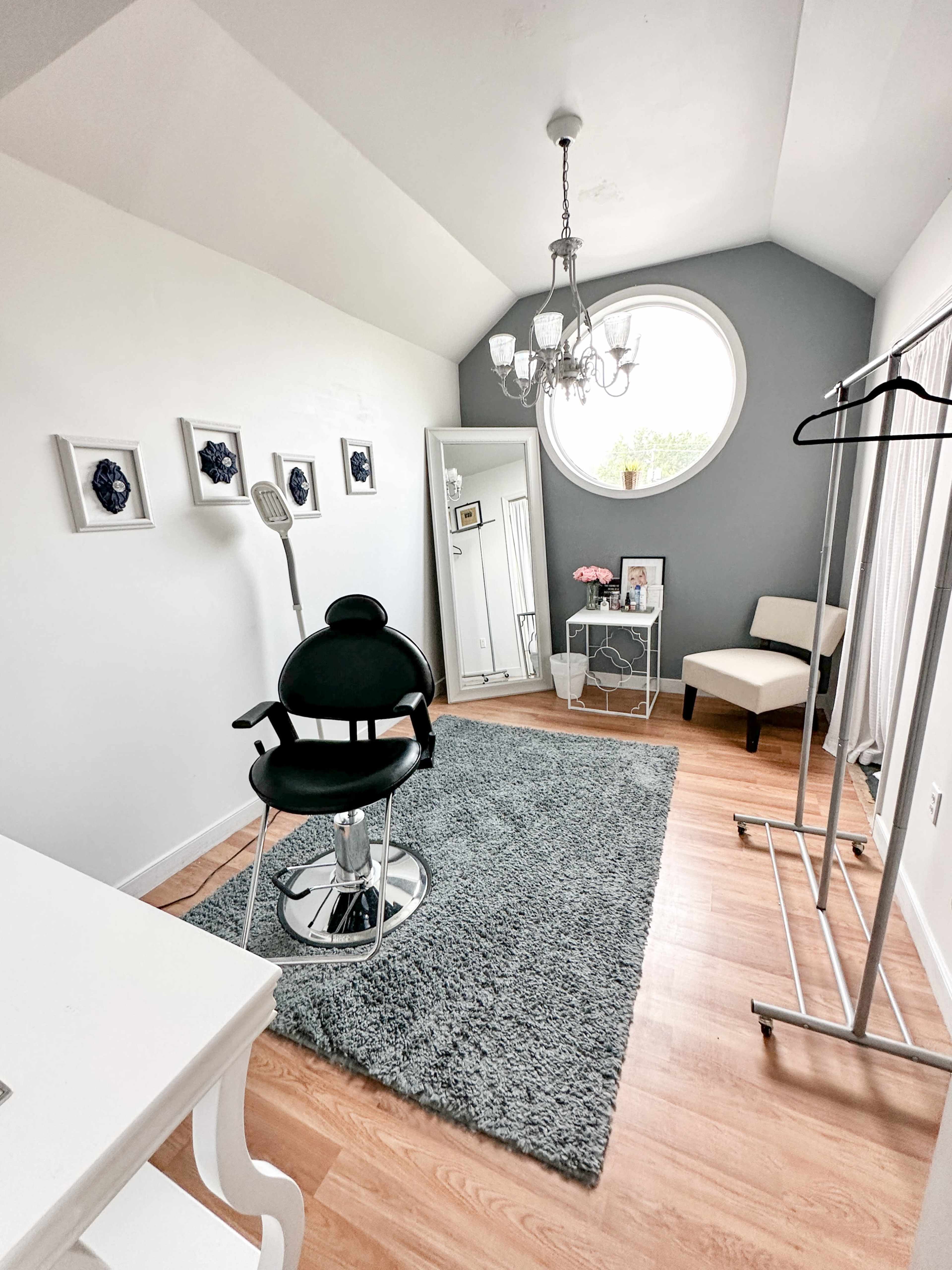 A well-lit room featuring a salon chair, a small table with a photo, a clothing rack, and framed artwork on the walls.