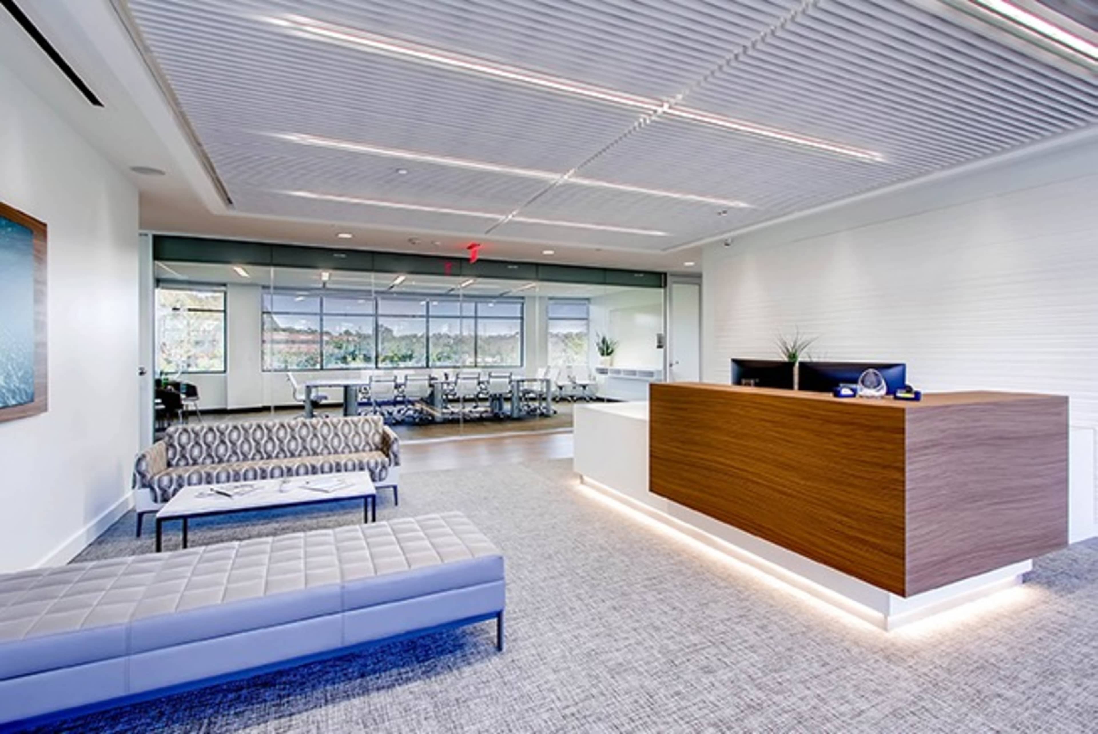 The image shows a modern office reception area with a wooden reception desk, seating options, and a conference room visible in the background.