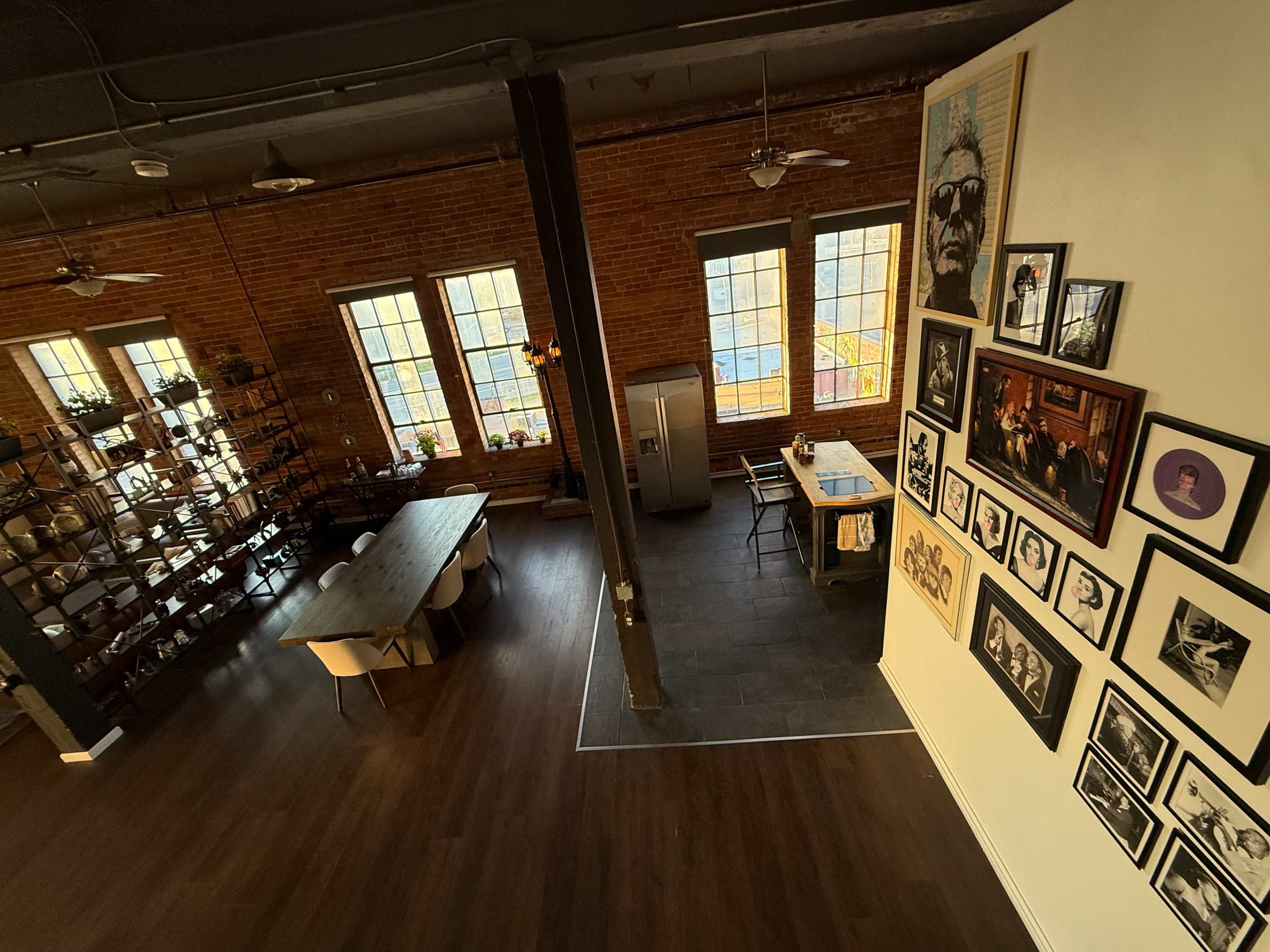 The image shows a spacious industrial-style kitchen and dining area with exposed brick walls, a large wooden table, and a gallery of framed artworks on one wall.