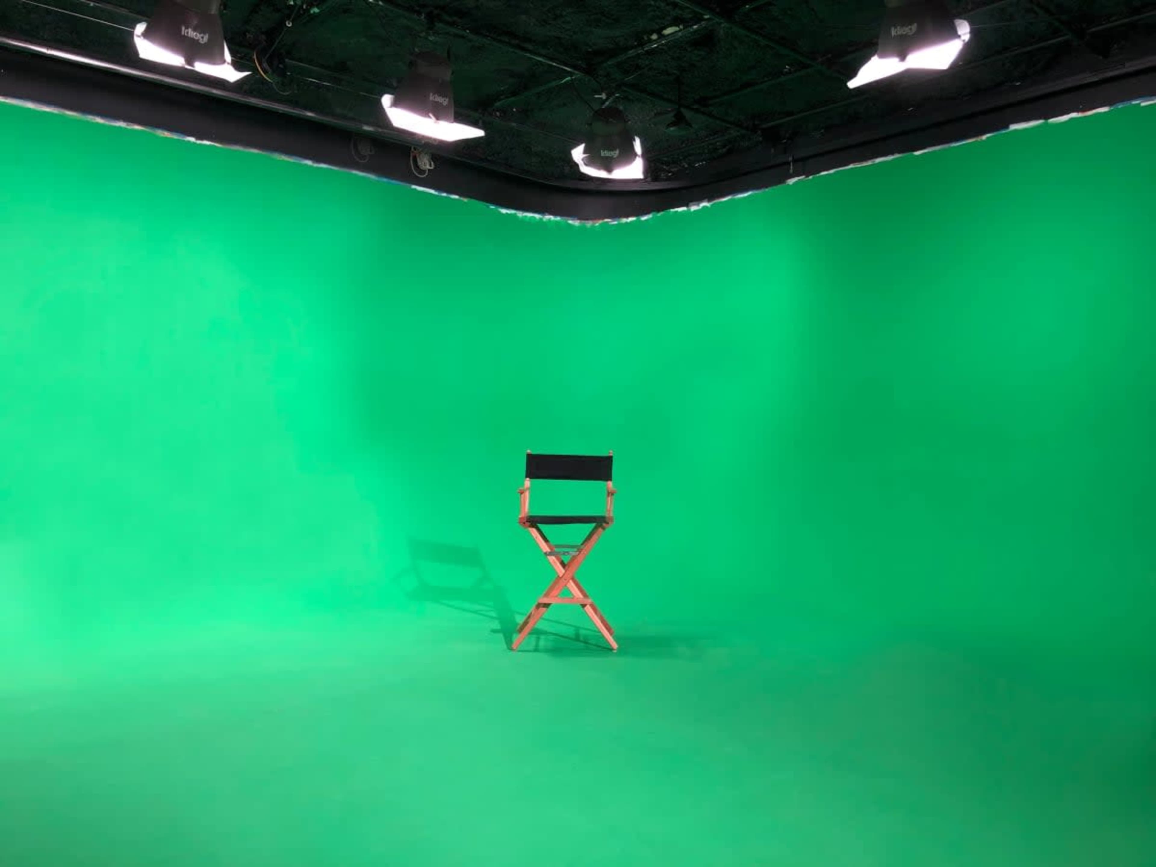 A director's chair is positioned in the center of a green screen studio with overhead lights illuminating the space.
