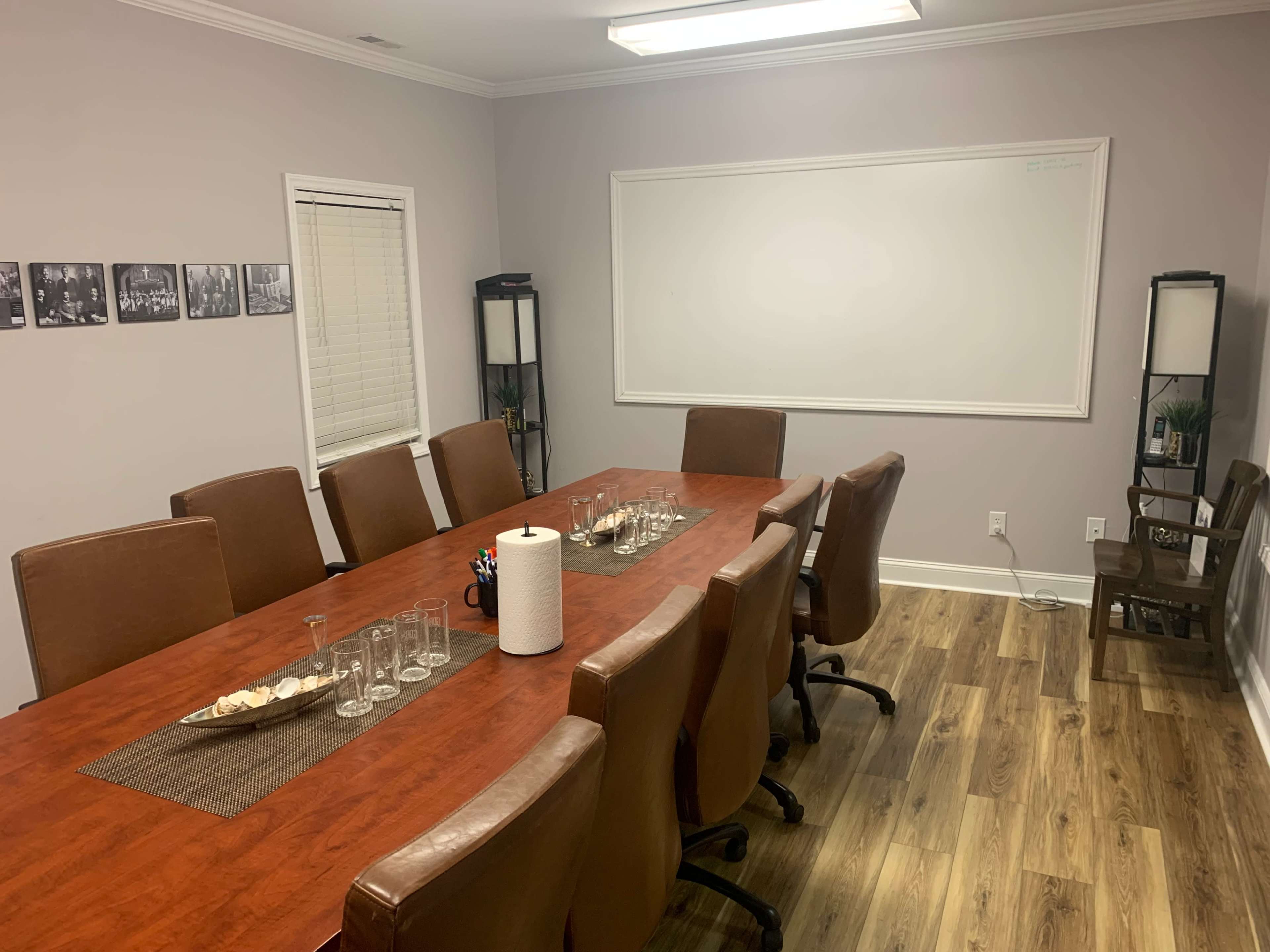 The image shows a conference room with a large wooden table surrounded by leather chairs, a whiteboard on the wall, and decorative elements like a tray with glasses and paper towels.