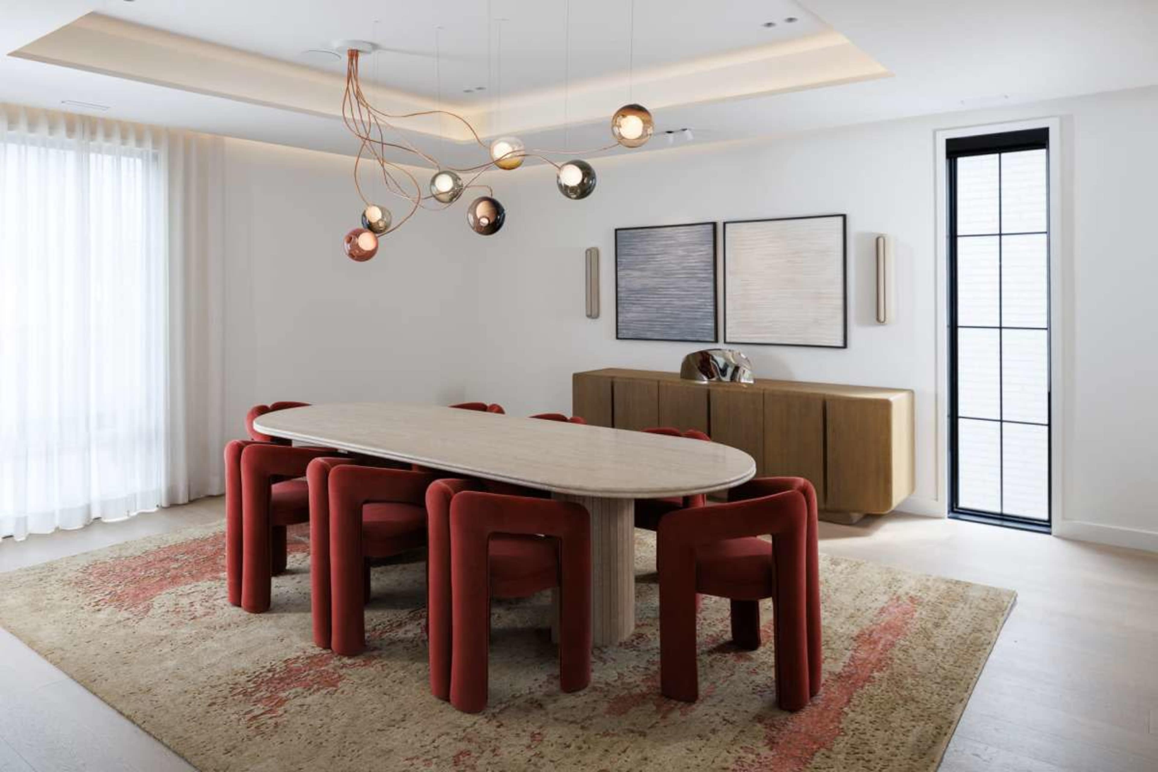 The image shows a modern dining room featuring a long oval table surrounded by red upholstered chairs, with decorative pendant lights above and a sideboard against the wall.