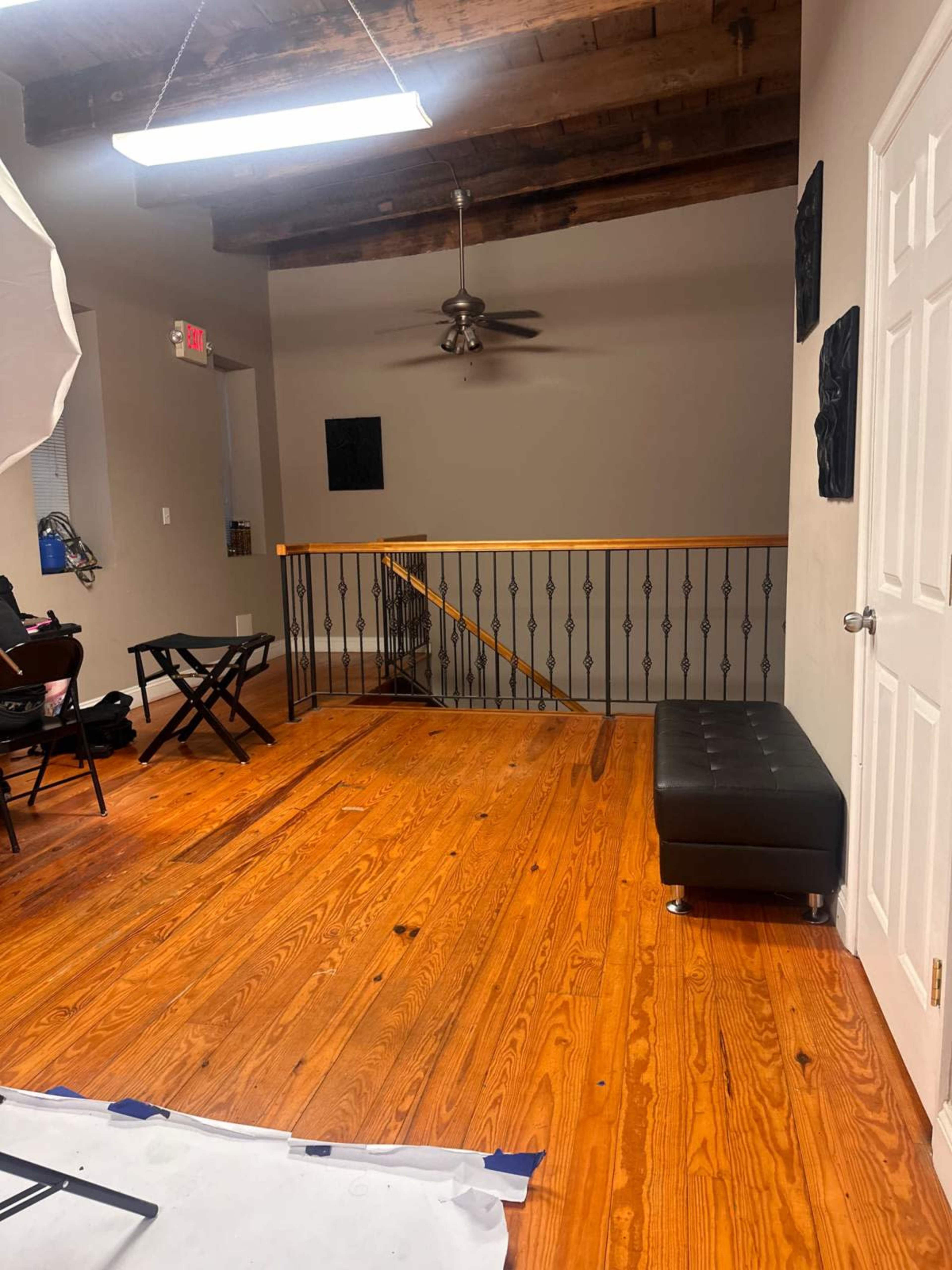 The image depicts a spacious room with wooden floors, a ceiling fan, a staircase railing, and a black bench against a wall.