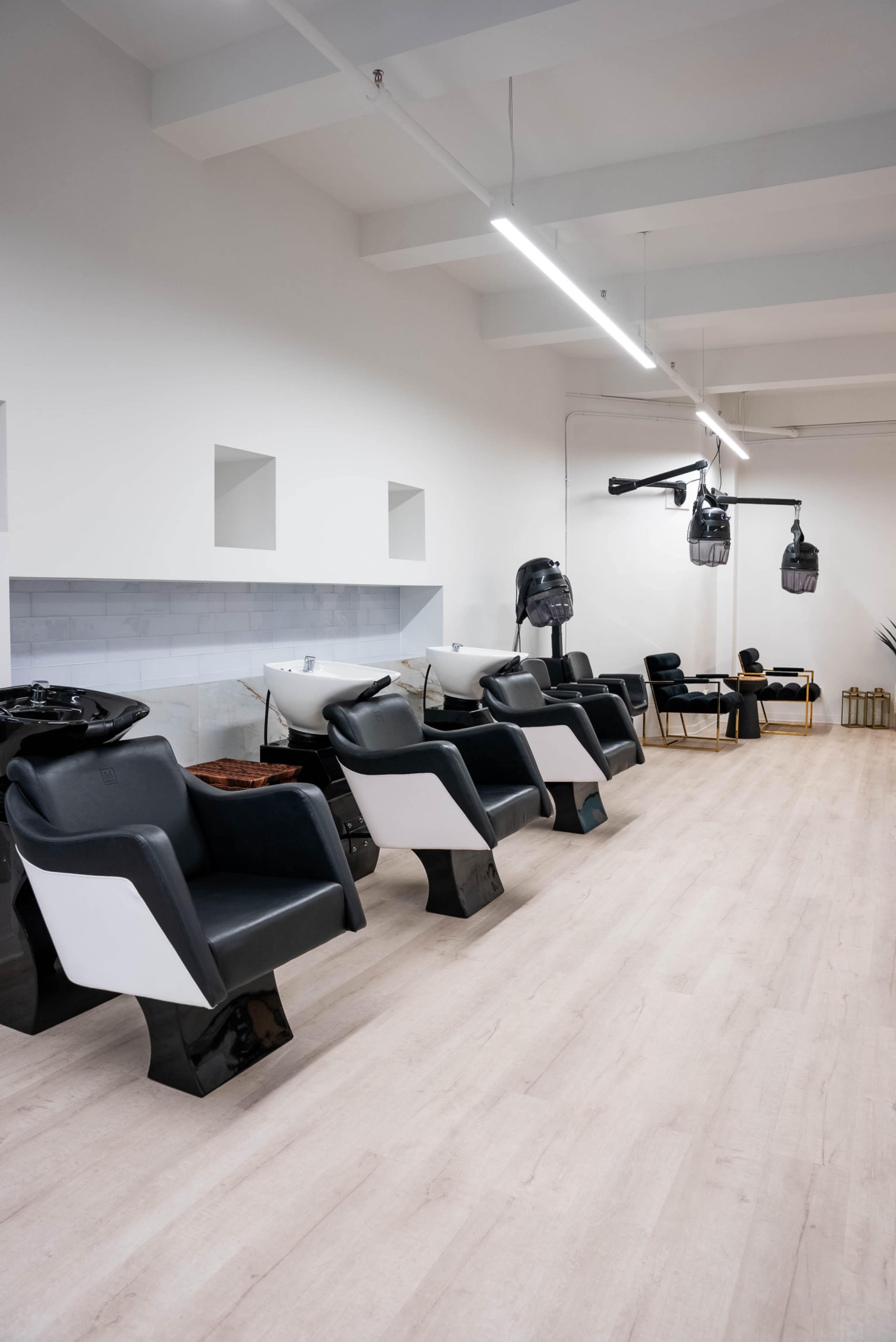 A modern hair salon interior with black and white styling chairs, wash stations, and hairdryers suspended from the ceiling.