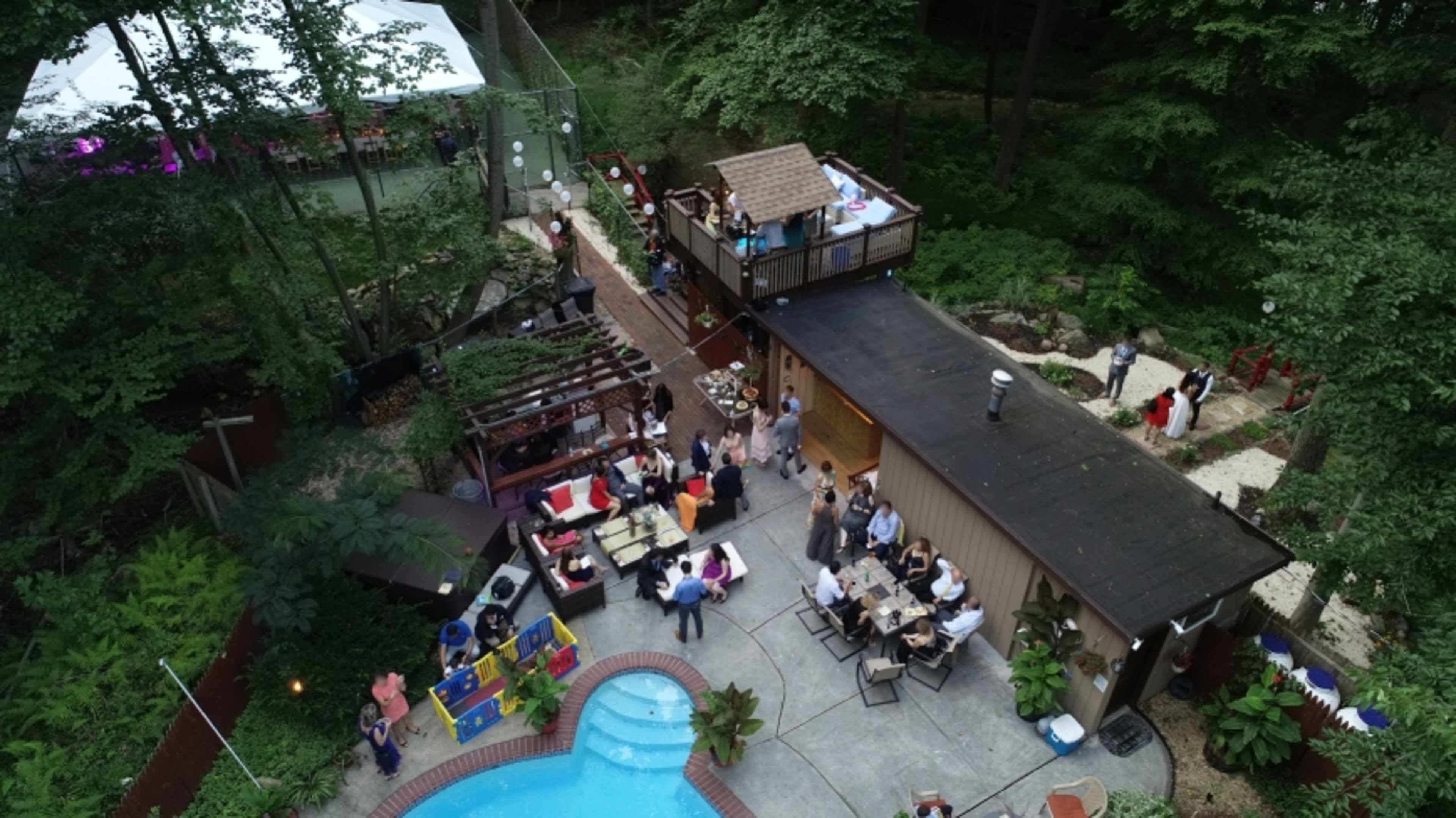 An outdoor gathering takes place around a pool, with guests socializing near a house and a separate deck area surrounded by trees.