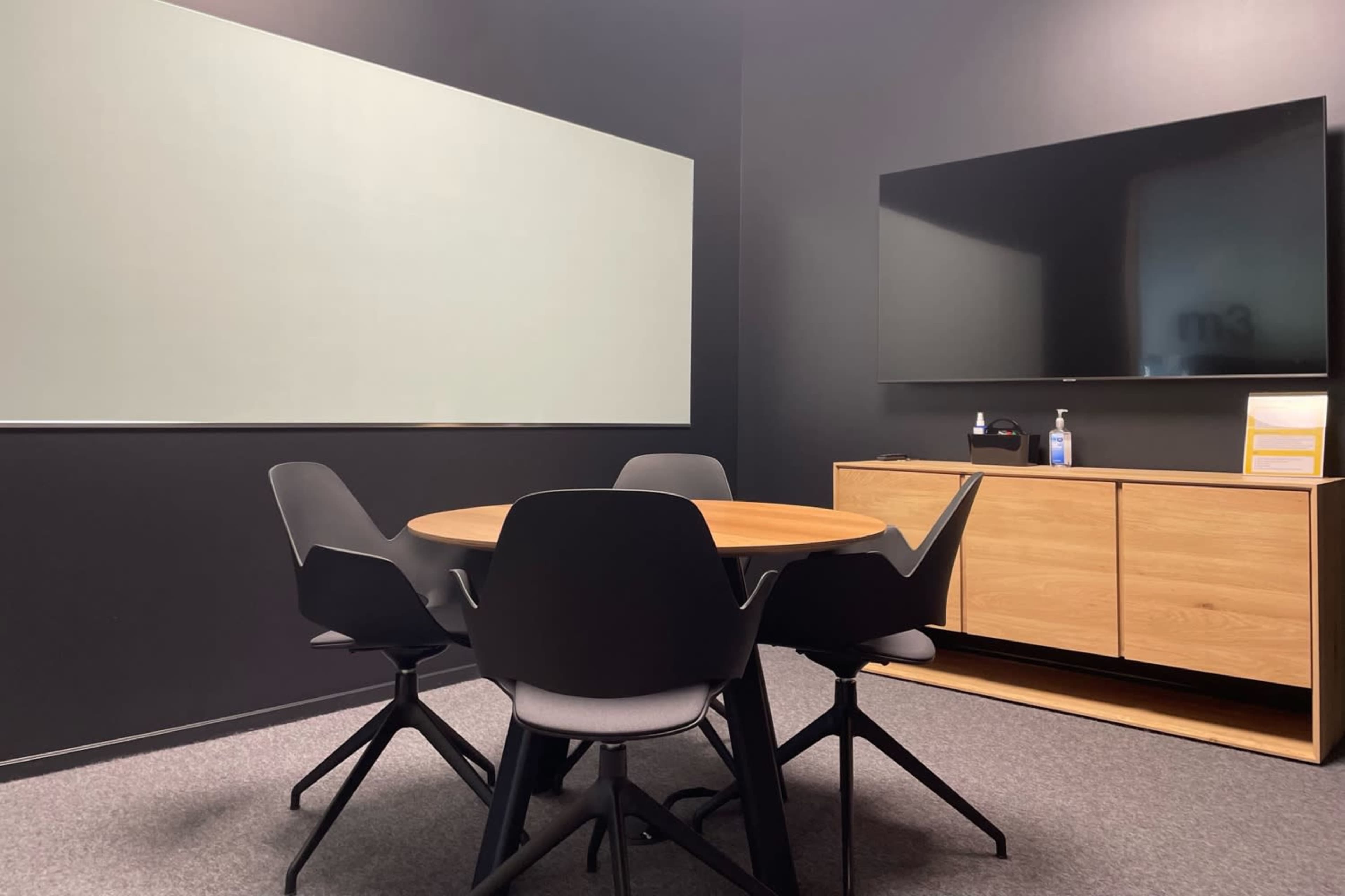 A small conference room featuring a round wooden table surrounded by black chairs, a whiteboard, and a wall-mounted television.