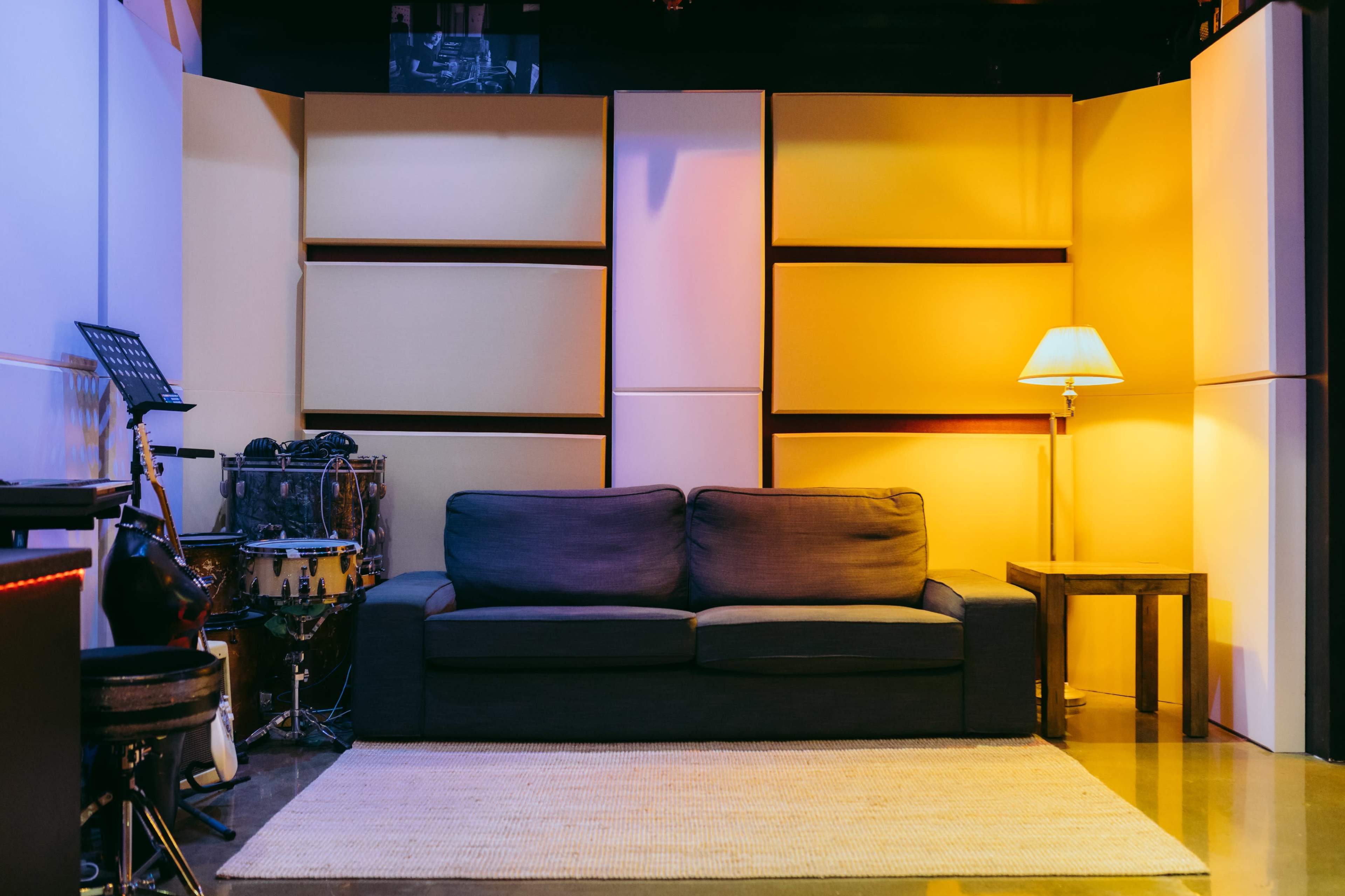 The image shows a cozy indoor setup with a dark sofa, a small wooden table, a floor lamp, and a musical instrument setup in a warmly lit space.