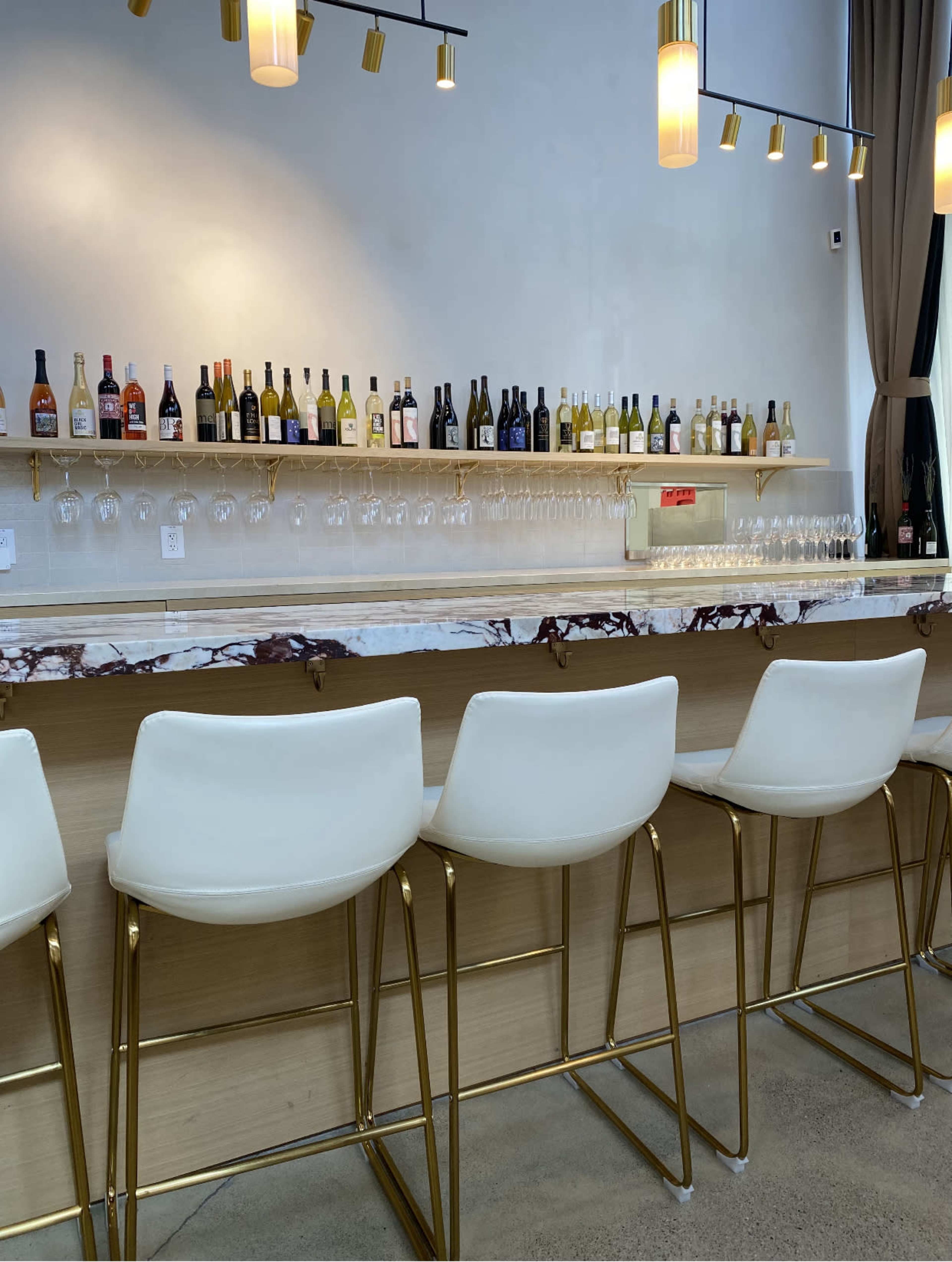 A modern bar area features a marble countertop, white stools with gold legs, and a shelf lined with various bottles of liquor and wine.
