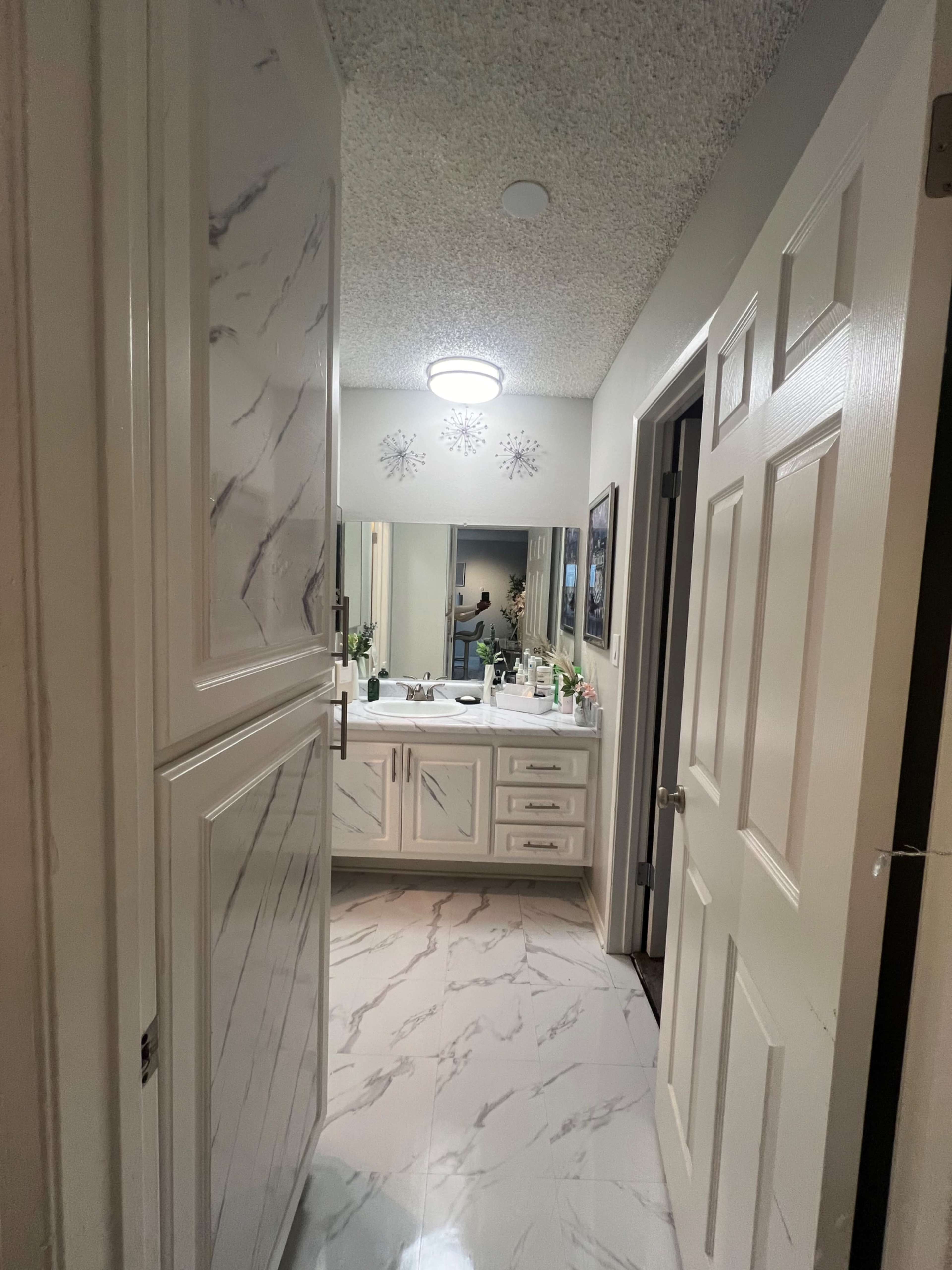 A bathroom with a hallway entrance, featuring a double vanity, mirror, and marble-like flooring.