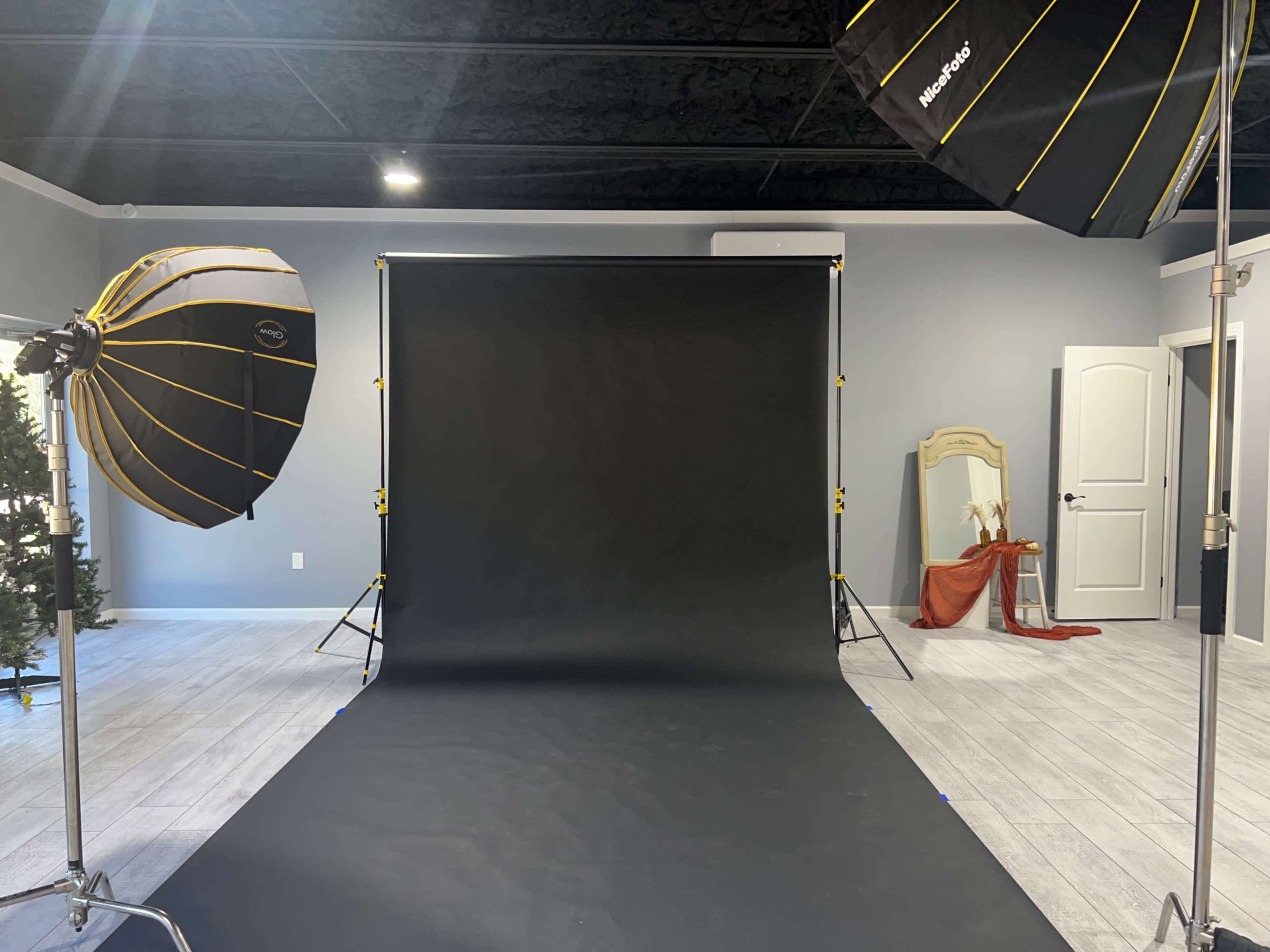The image shows a photography studio with a black backdrop, two lighting umbrellas on either side, and a decorated area with a mirror and a draped fabric in the corner.