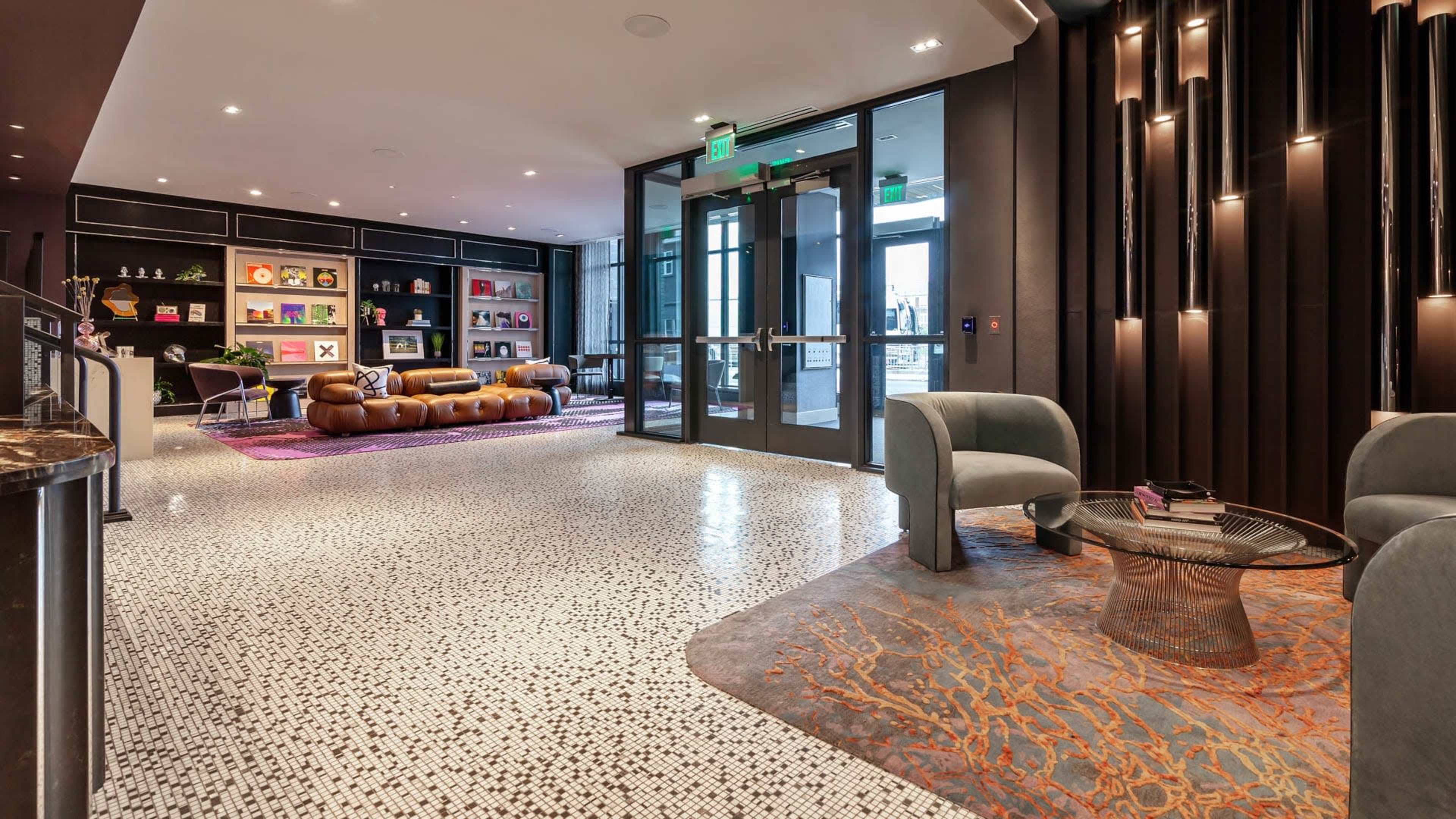 A modern lobby features a seating area with brown leather sofas, distinct rugs, and decorative wall shelves, leading to glass doors.