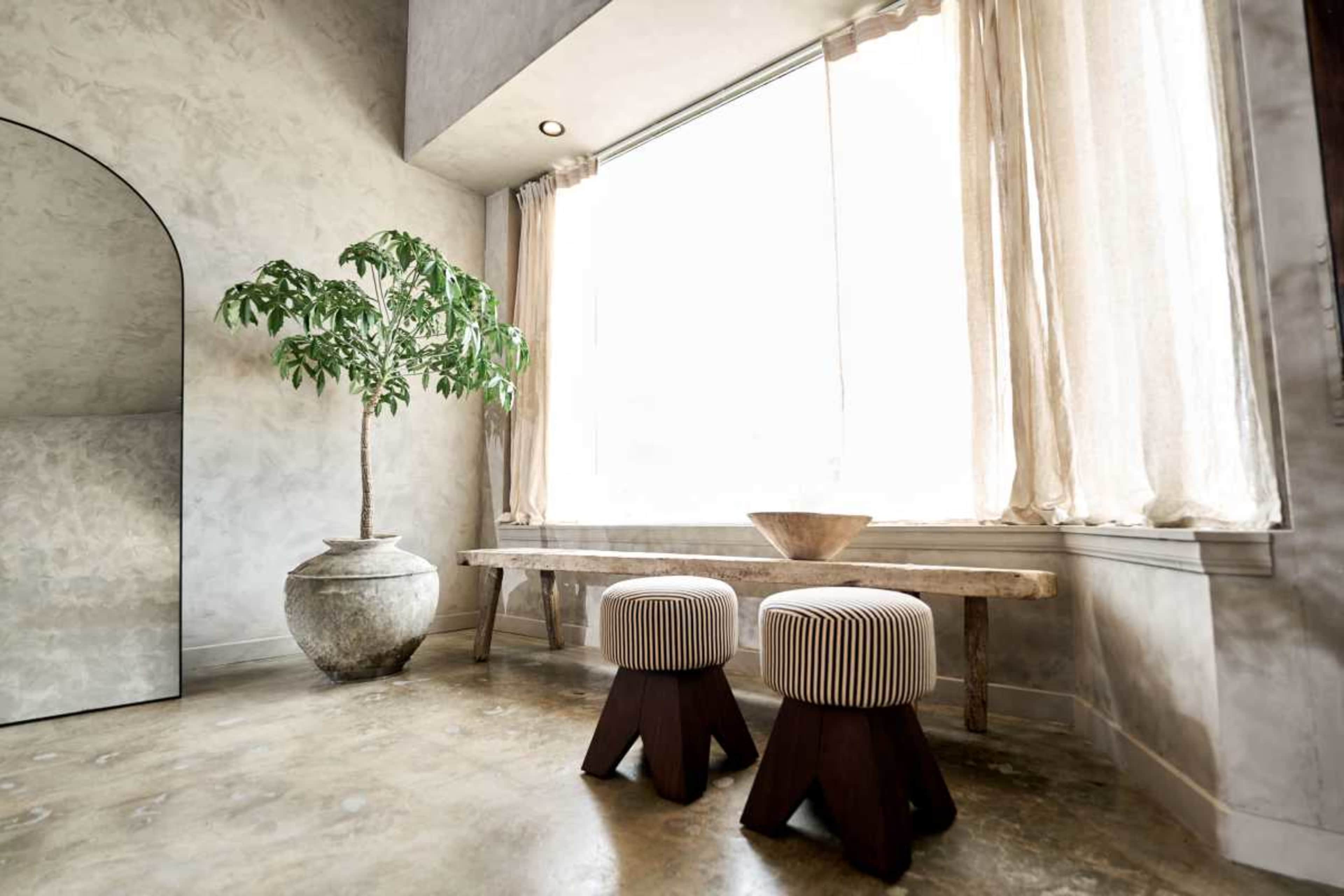 The image shows a minimalist interior with a large window, a potted plant, a wooden bench, and two striped ottomans.