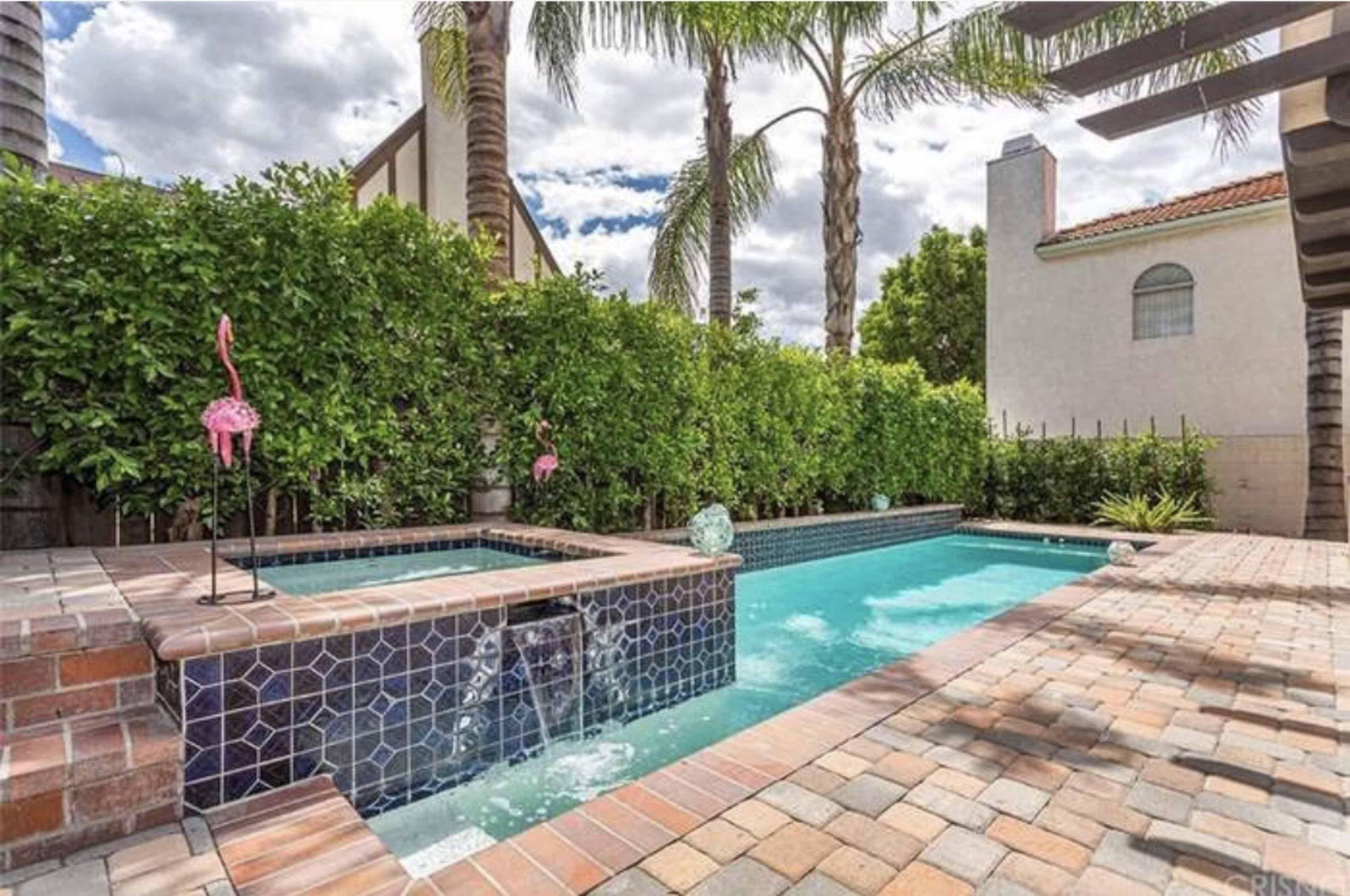 A pool area with a spa, surrounded by greenery and palm trees, featuring decorative flamingos and brick paving.