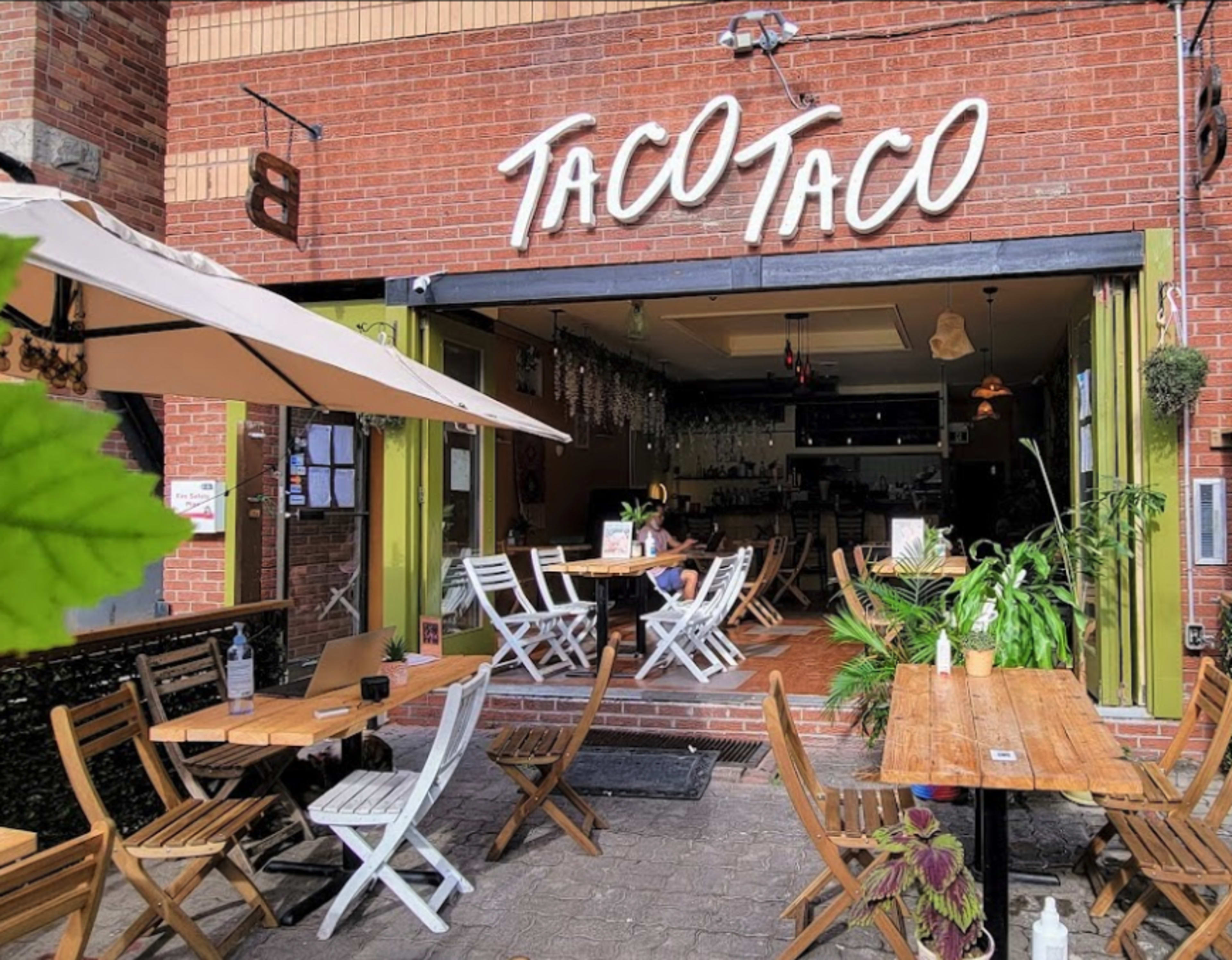 The exterior of a taco restaurant named "Taco Taco," featuring outdoor seating with wooden tables and chairs.