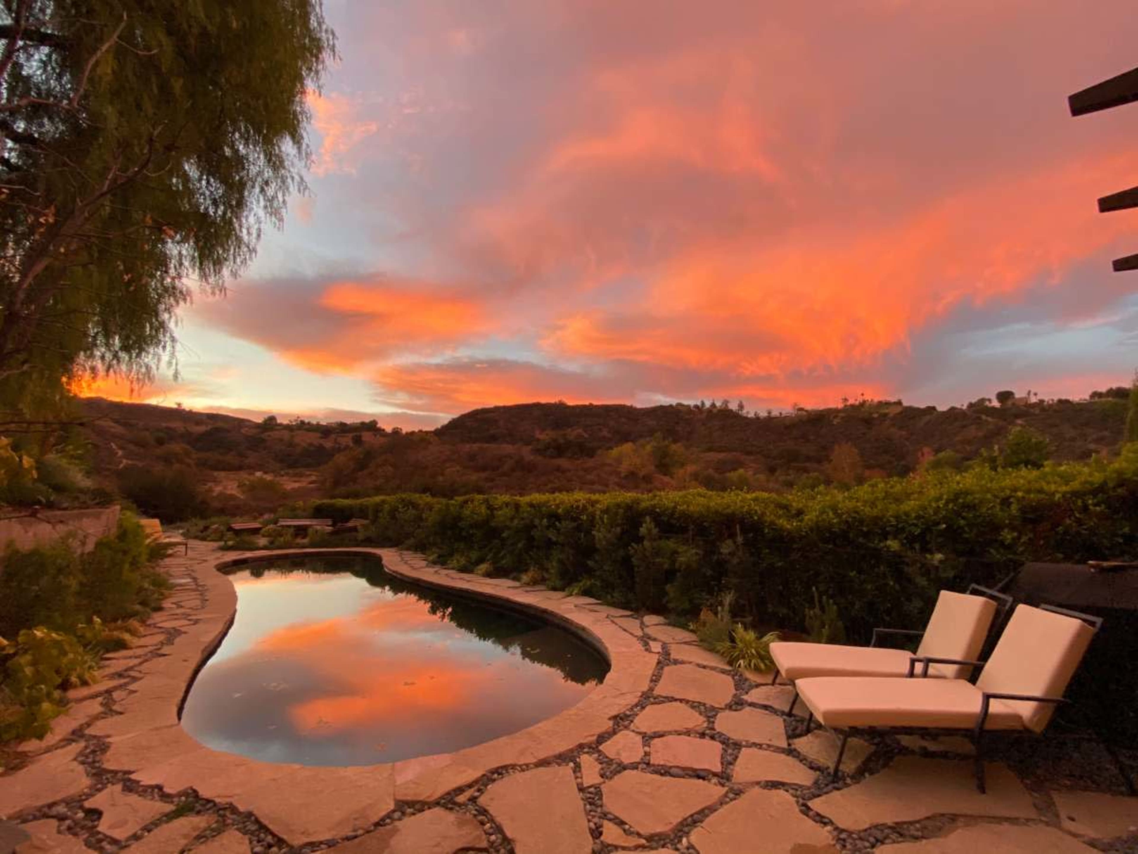 A tranquil pool reflects a vibrant sunset over rolling hills, framed by vegetation and stone landscaping.
