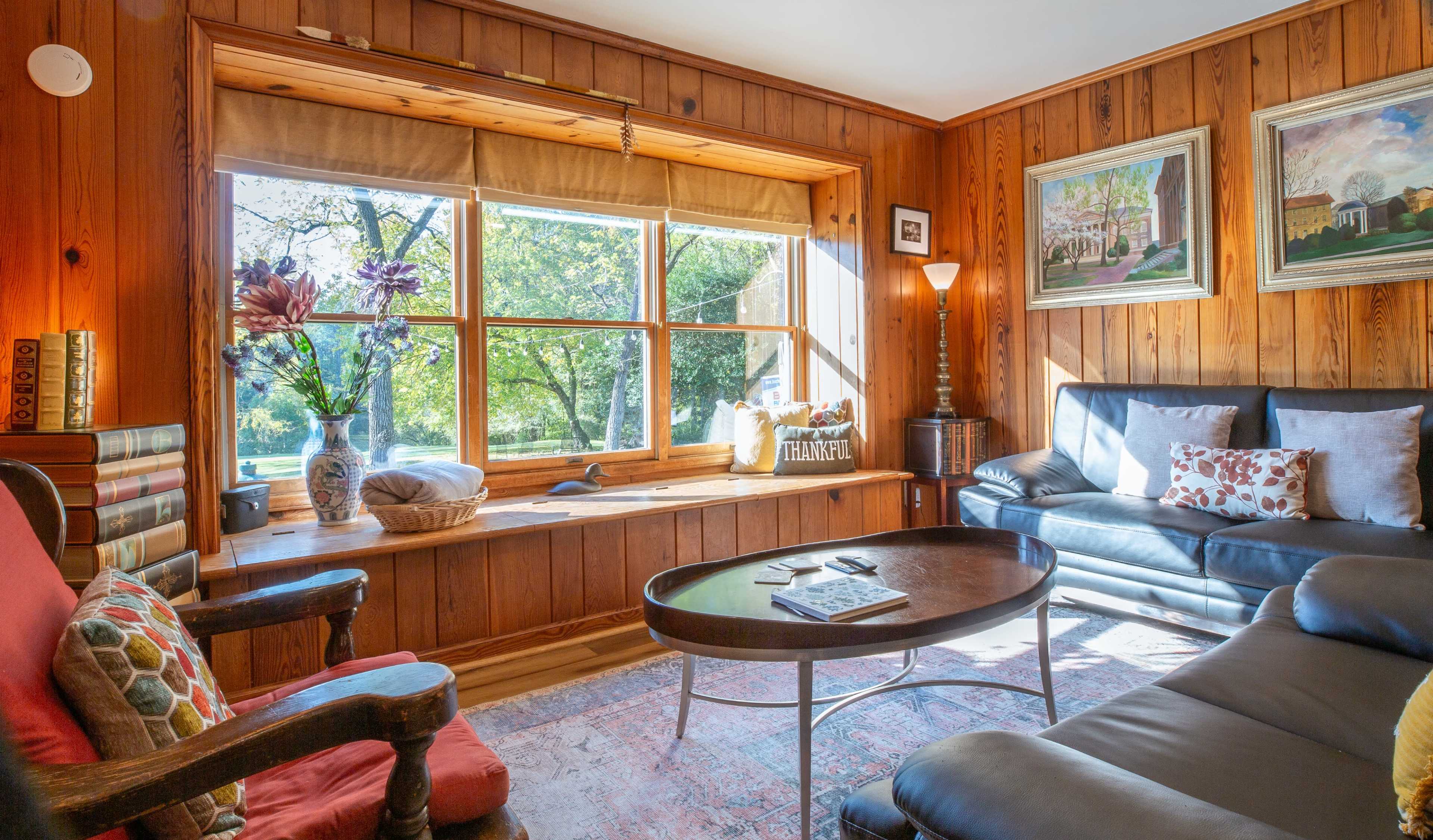 The image shows a cozy living room with wooden walls, featuring a large window that lets in natural light, a coffee table in the center, and comfortable seating arrangements.