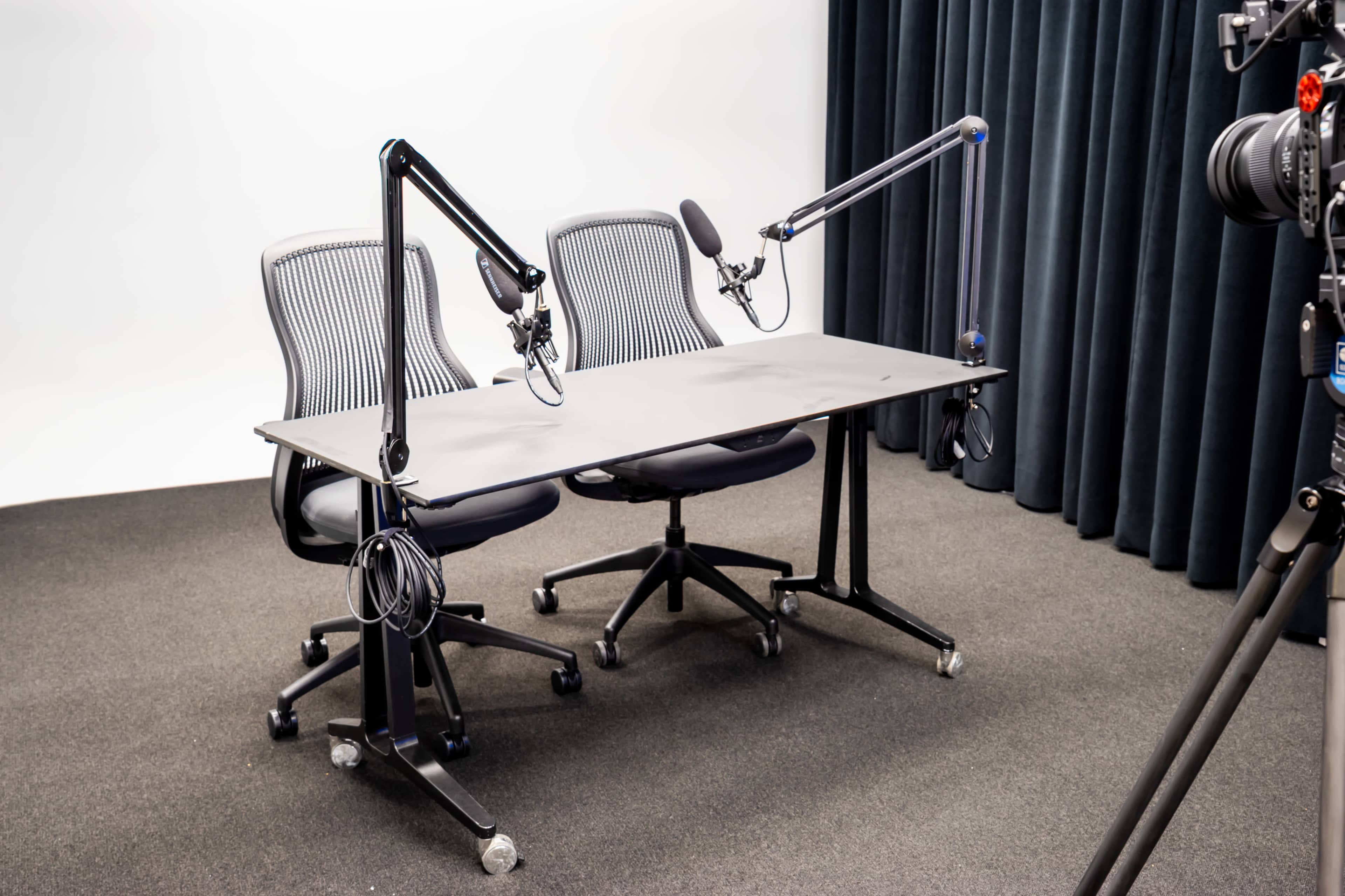 The image shows a recording setup with two ergonomic chairs, a black table, and two adjustable microphones positioned overhead.