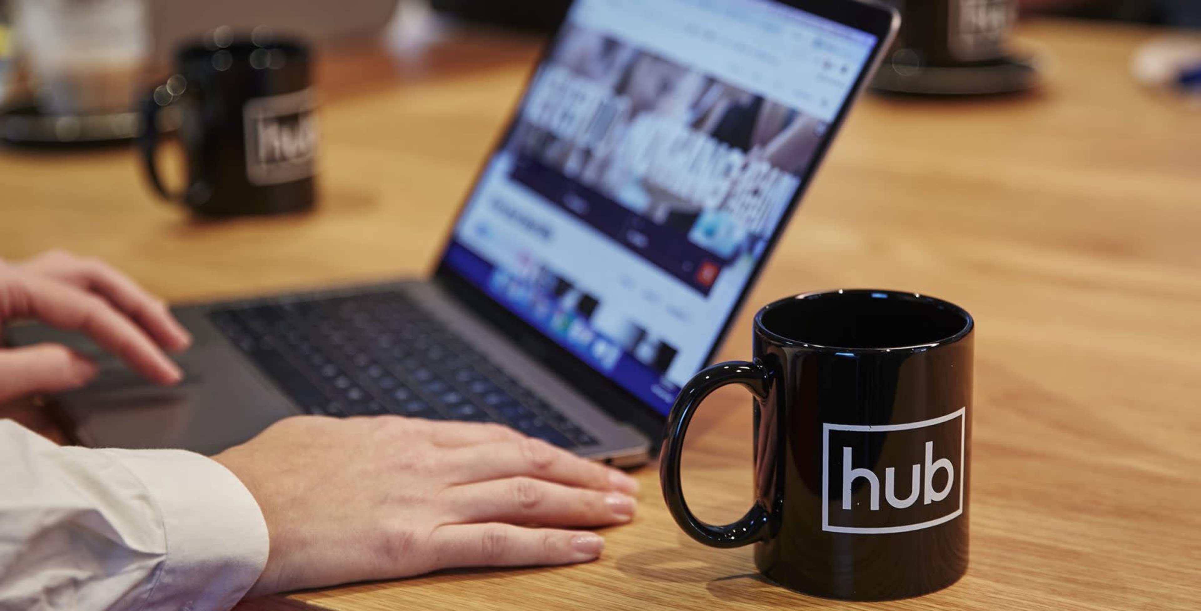 A person uses a laptop on a wooden table with a black coffee mug featuring the logo "hub" nearby.