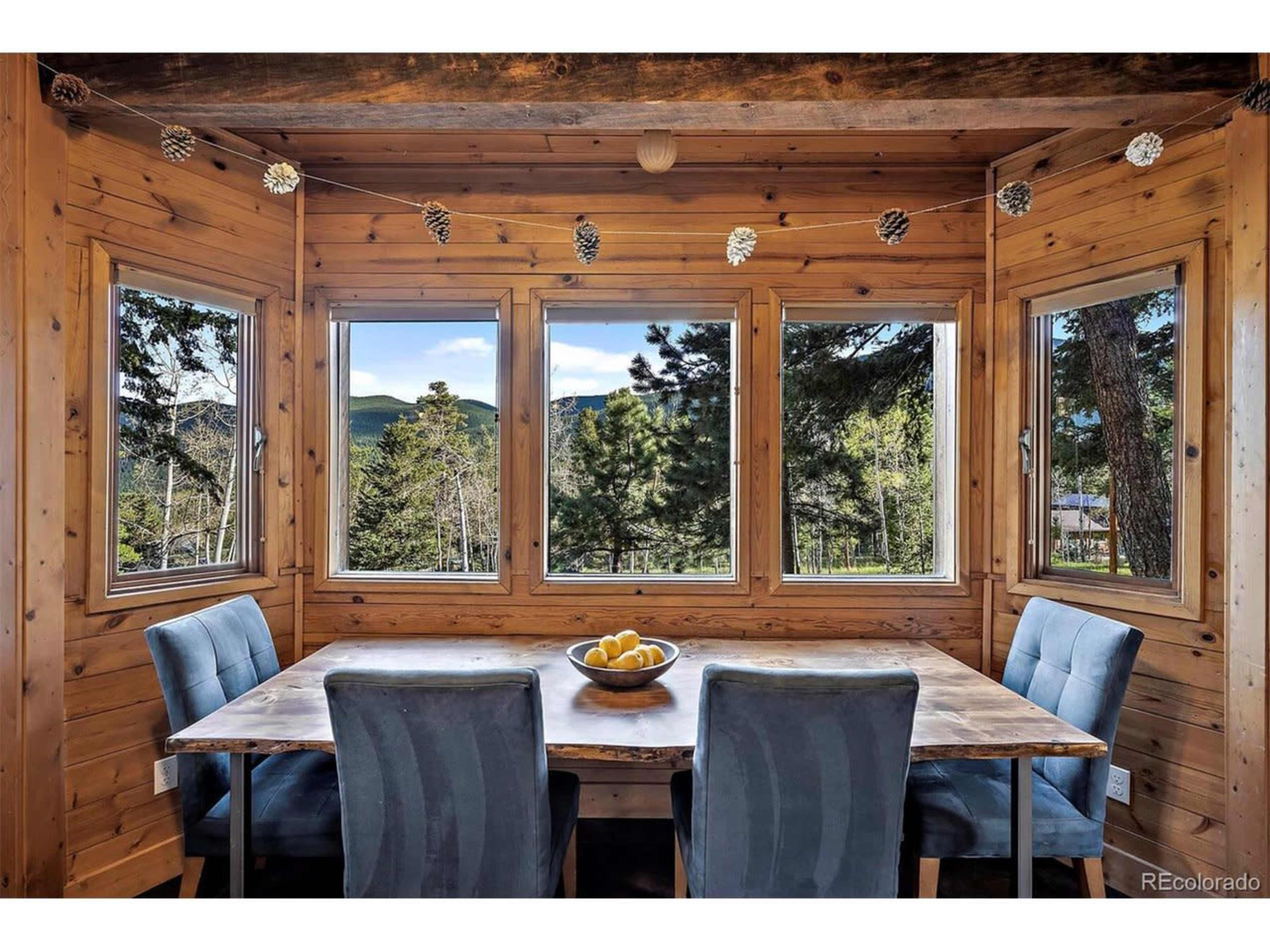 A wooden dining area features a large table surrounded by blue chairs, with large windows showing a view of trees and mountains.